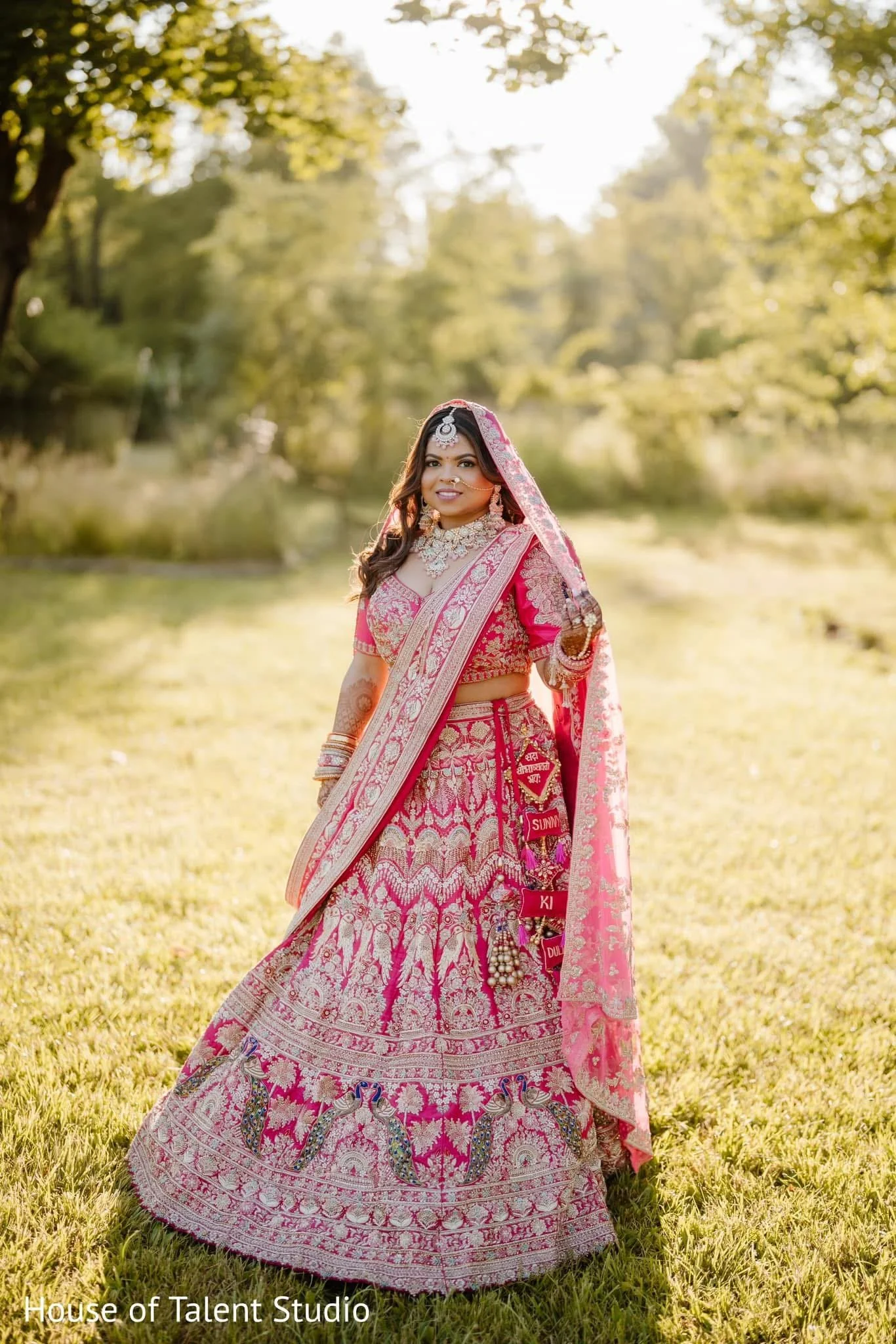 A woman in a vibrant pink traditional Indian bridal outfit standing in a lush outdoor setting with green trees and sunlight.
