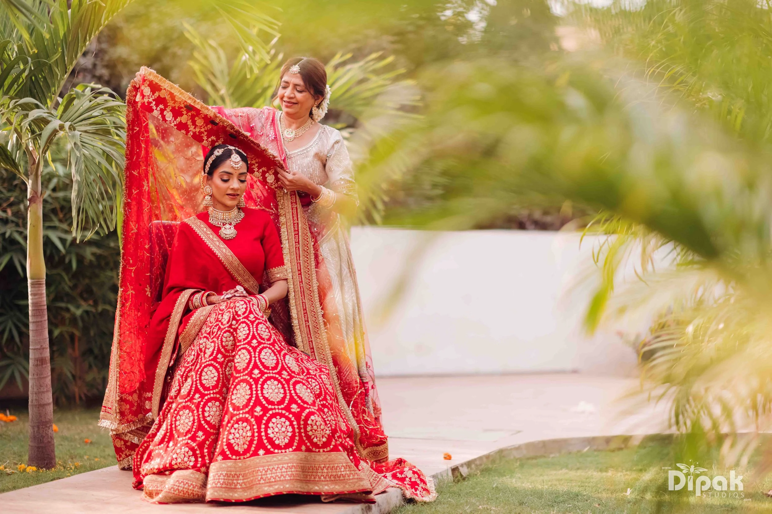 A woman dressed in a red traditional Indian saree with intricate gold embroidery, sitting outdoors on a small platform, with another woman standing behind her, adjusting her saree or jewelry, surrounded by greenery and palm trees.