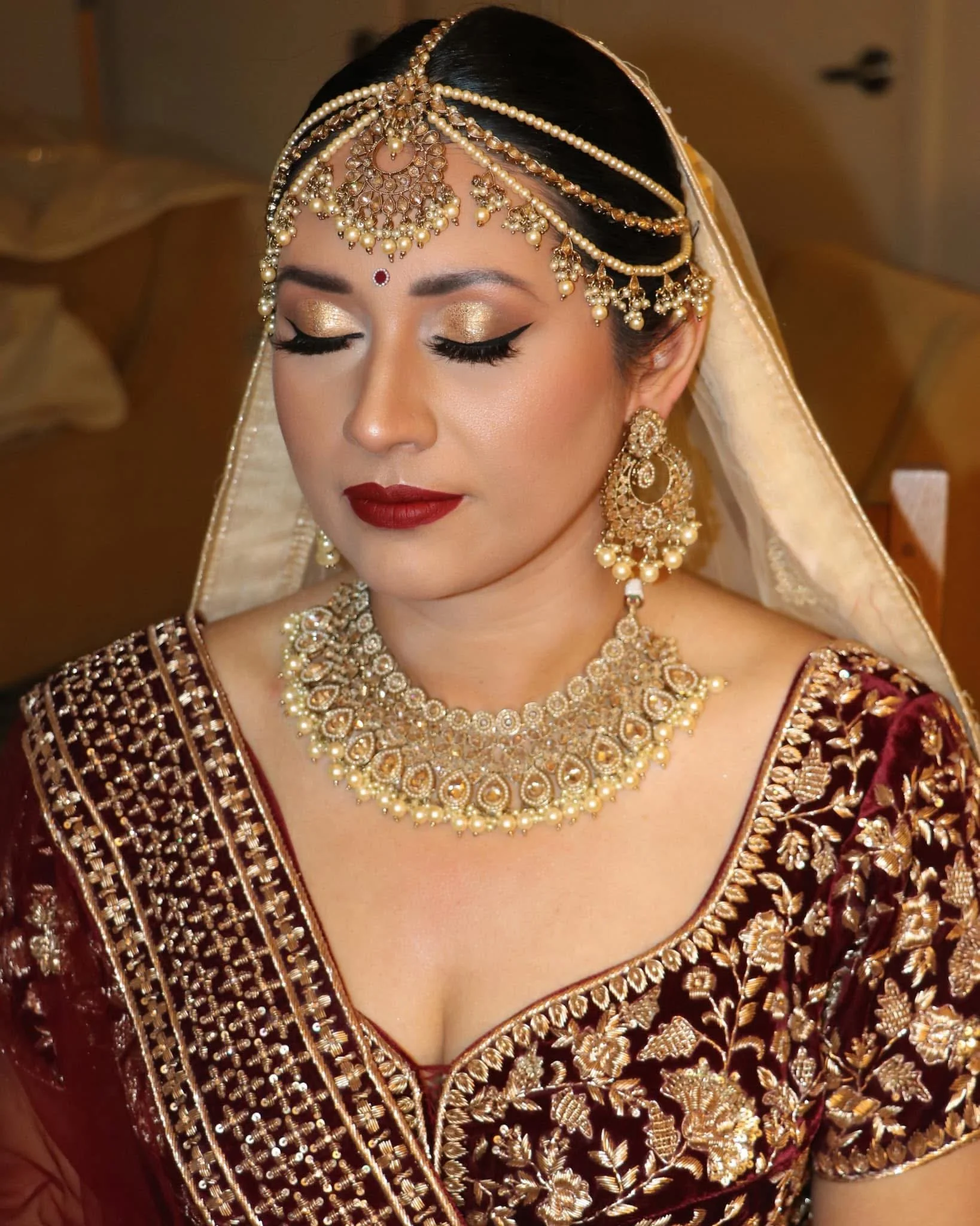A woman in traditional Indian bridal attire with gold jewelry, including a headpiece, necklace, earrings, and a deep red and gold embroidered outfit.
