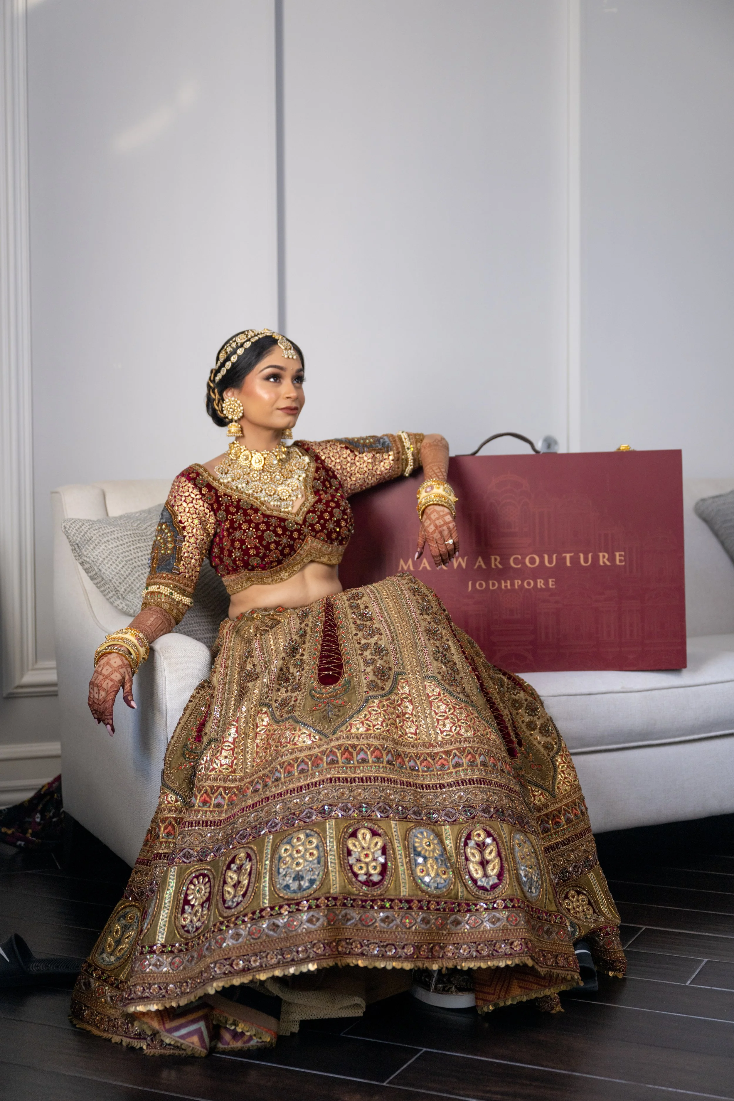A woman wearing traditional Indian bridal attire, including a heavily embroidered lehenga, jewelry, and henna on her hands, sitting on a white couch with a maroon bag that reads "MOTWARCOUTURE JODHPUR" beside her.