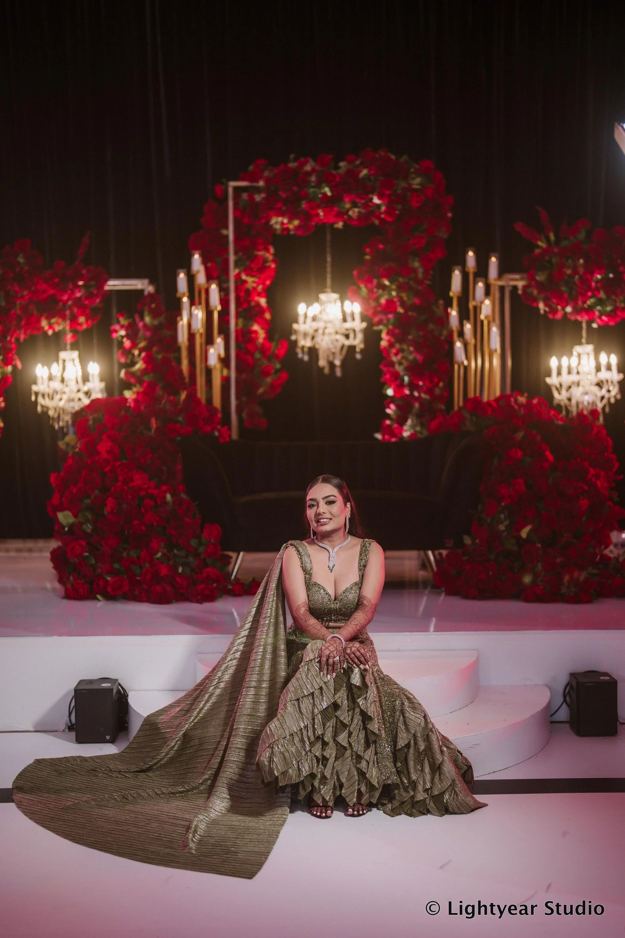 A woman in a gold and brown traditional Indian dress sitting on stage with a large gold fabric train spread out in front of her, in front of a decorated stage with red roses, chandeliers, and candelabra.