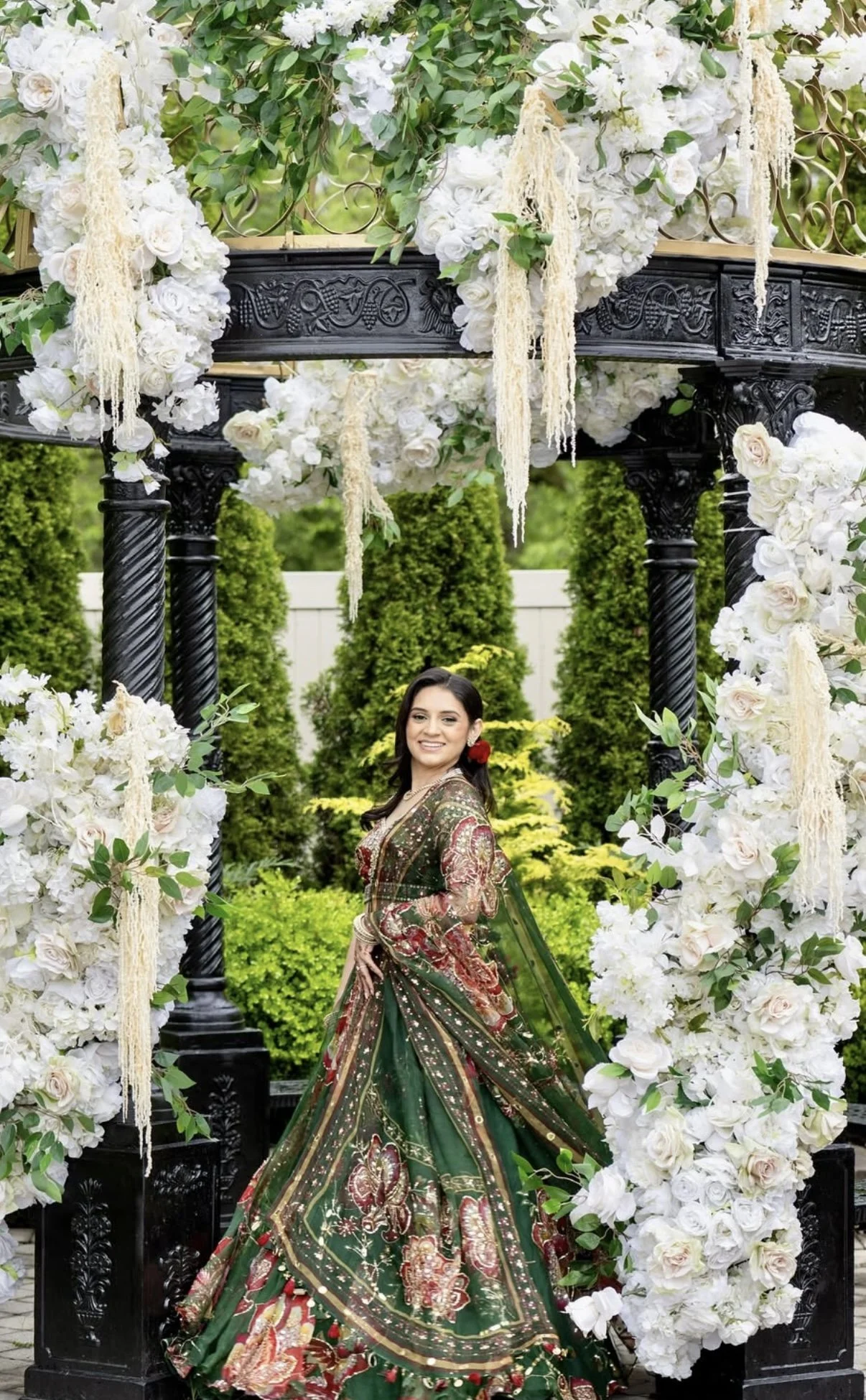 A woman in a colorful traditional Indian saree stands under a decorative arch decorated with white flowers and greenery, with a garden background.