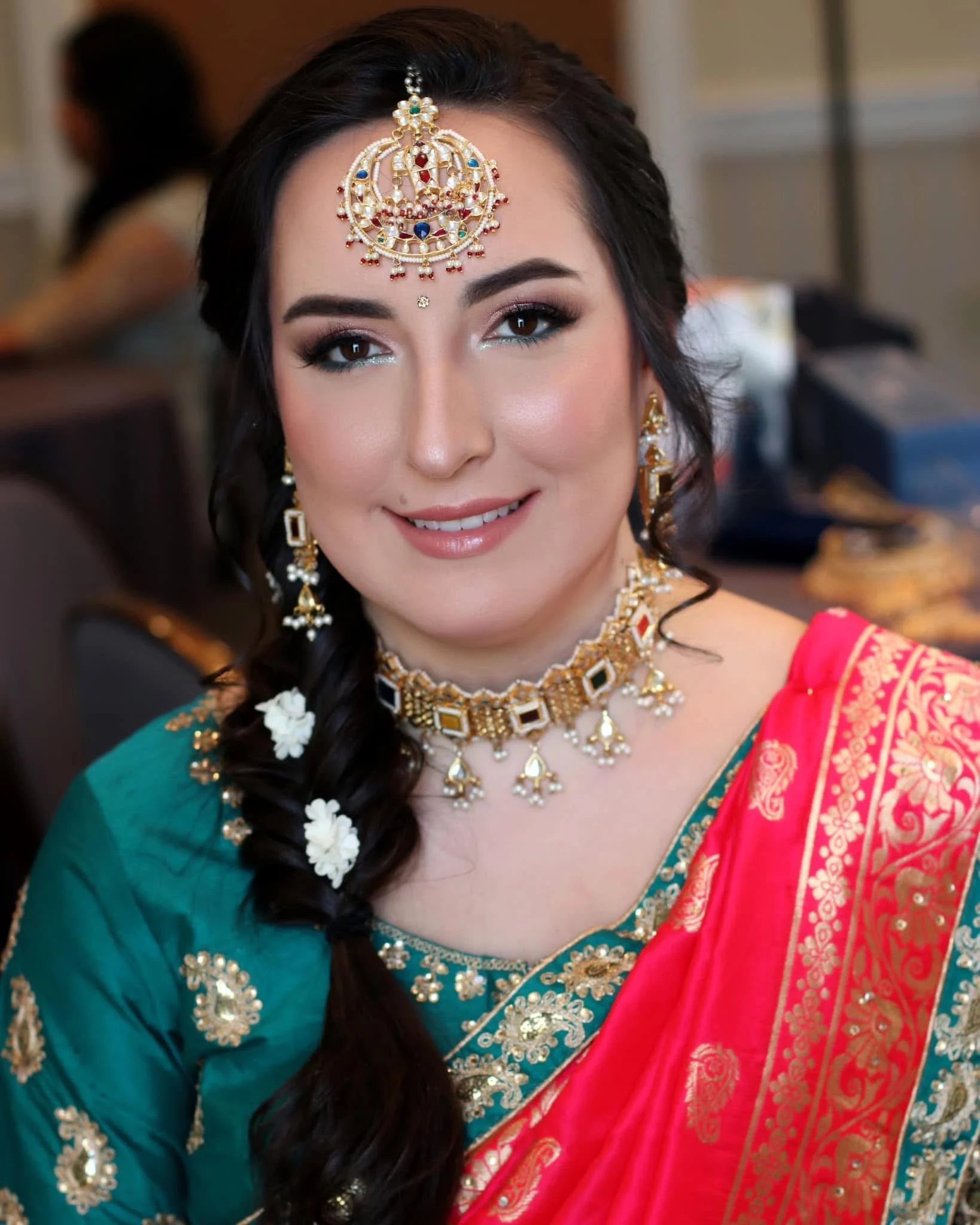 A woman dressed in traditional Indian attire, wearing jewelry including a maang tikka on her forehead, earrings, necklace, and bangles, with her hair styled in a braid decorated with white flowers.