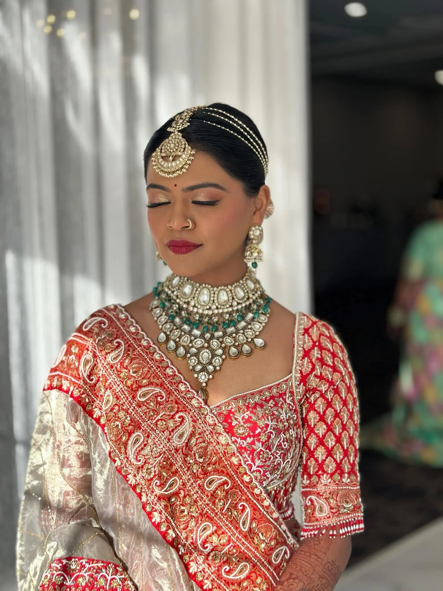 A woman dressed in traditional Indian attire with intricate jewelry, including a large necklace, earrings, a maang tikka, and bangles. She has her eyes closed and is wearing makeup, with a bindi on her forehead and a red and gold embroidered saree.