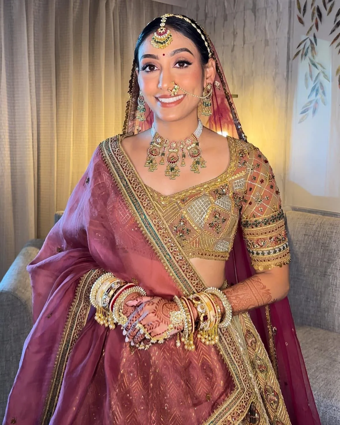 A woman dressed in traditional Indian bridal attire, wearing a maroon and gold embroidered saree with intricate jewelry, including a necklace, earrings, nose ring, bangles, and a headpiece, smiling with henna designs on her hands, standing indoors wi