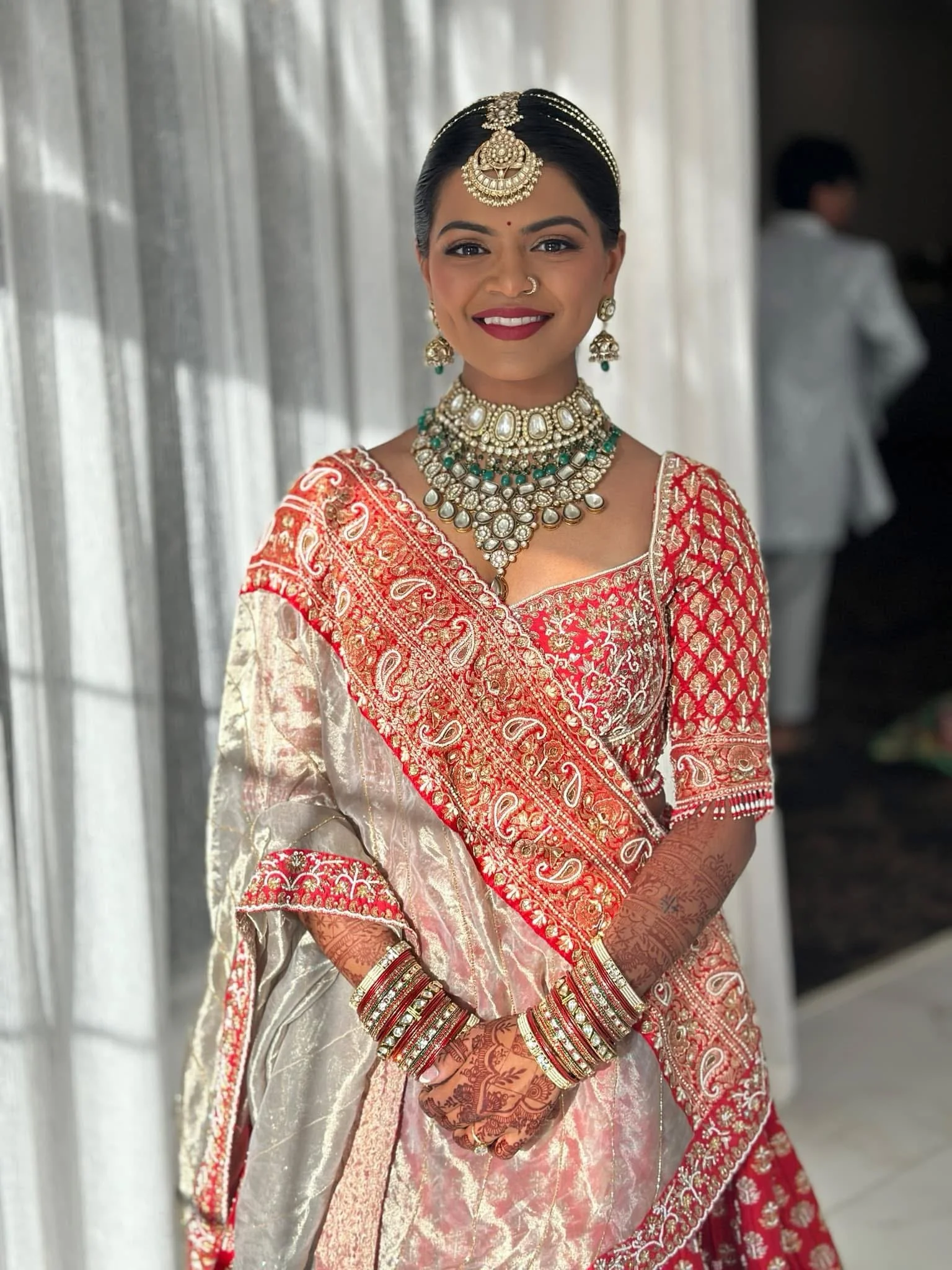 A woman dressed in traditional Indian attire, wearing a red and gold embroidered saree, with elaborate jewelry including necklaces, earrings, a maang tikka, and bangles. She has henna designs on her hands and is smiling.