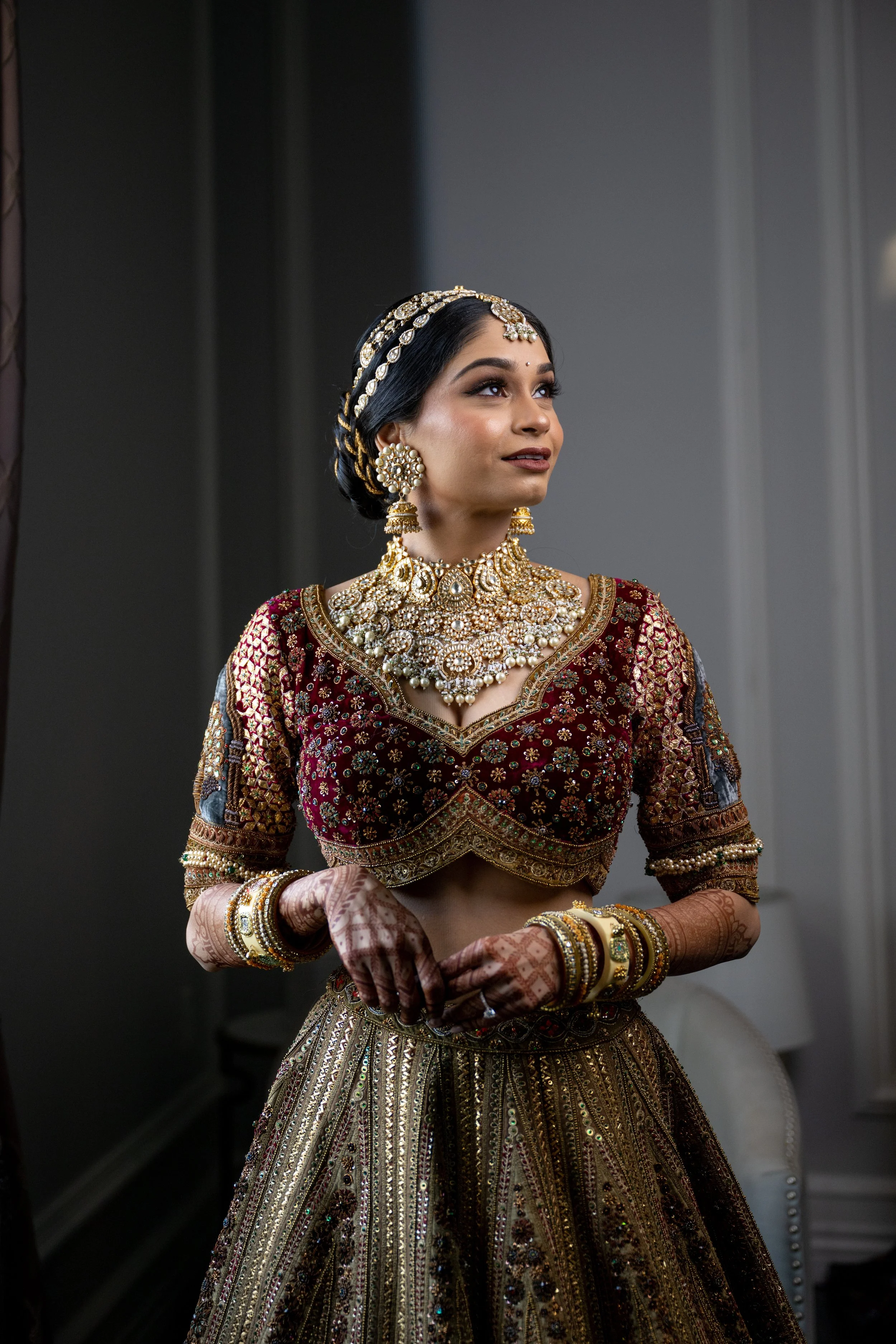 A woman dressed in traditional Indian bridal attire, wearing a red and gold embroidered blouse and a heavily embellished gold skirt. She is adorned with intricate jewelry including a choker necklace, large earrings, a headpiece, bangles, and henna on