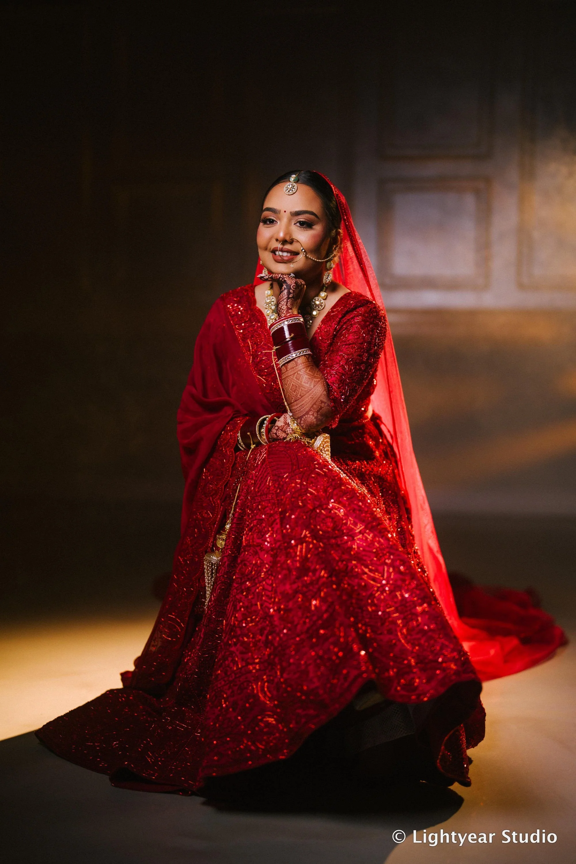 A woman dressed in a red traditional Indian outfit, seated on a dark surface, with a blurred background and soft lighting highlighting her attire and jewelry.