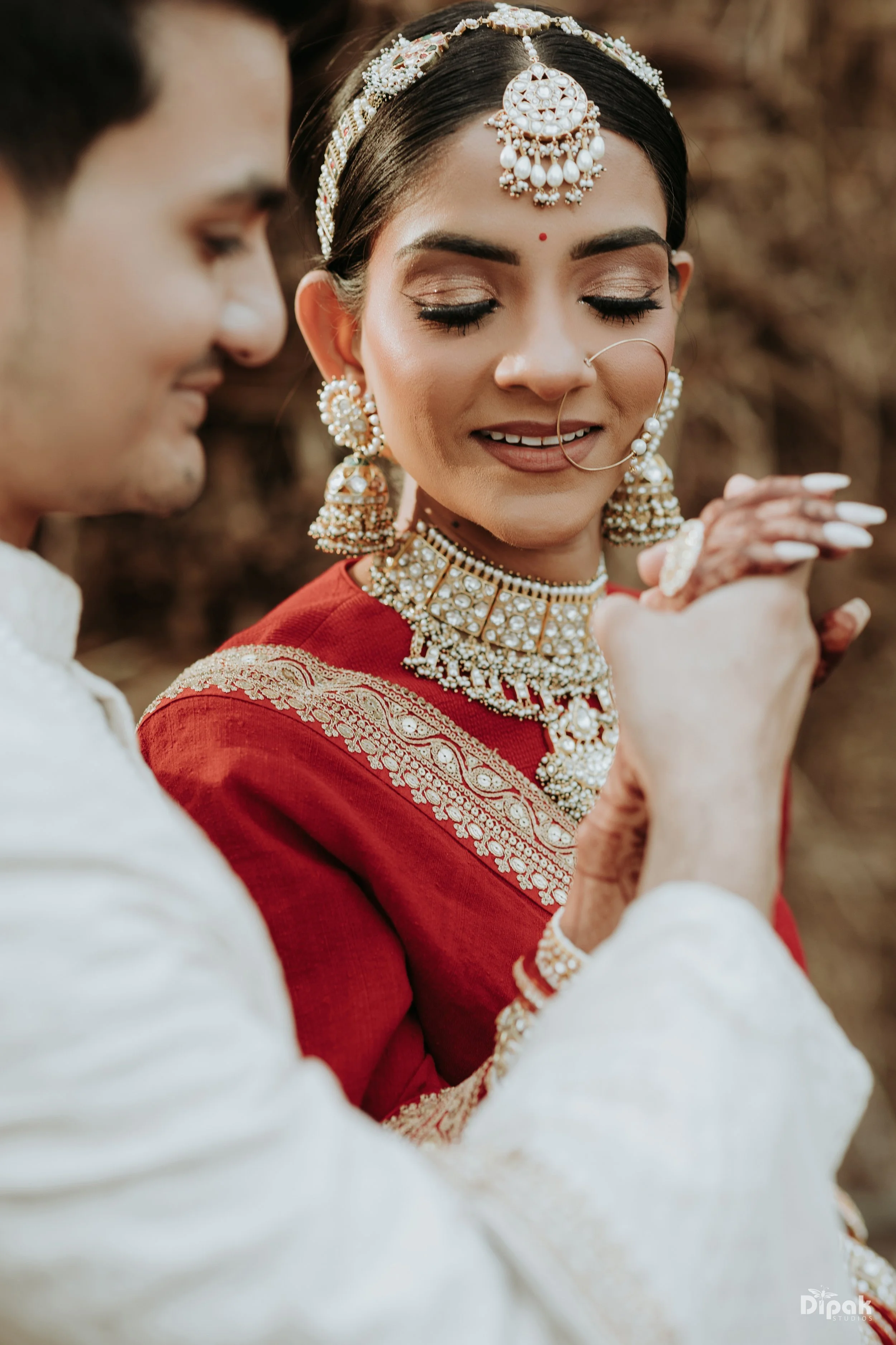 A woman dressed in traditional Indian attire with elaborate jewelry, including large earrings, a choker, forehead ornament, nose ring, and headpiece, smiling with her eyes closed while a man in white attire holds her hands in a close-up shot, likely 