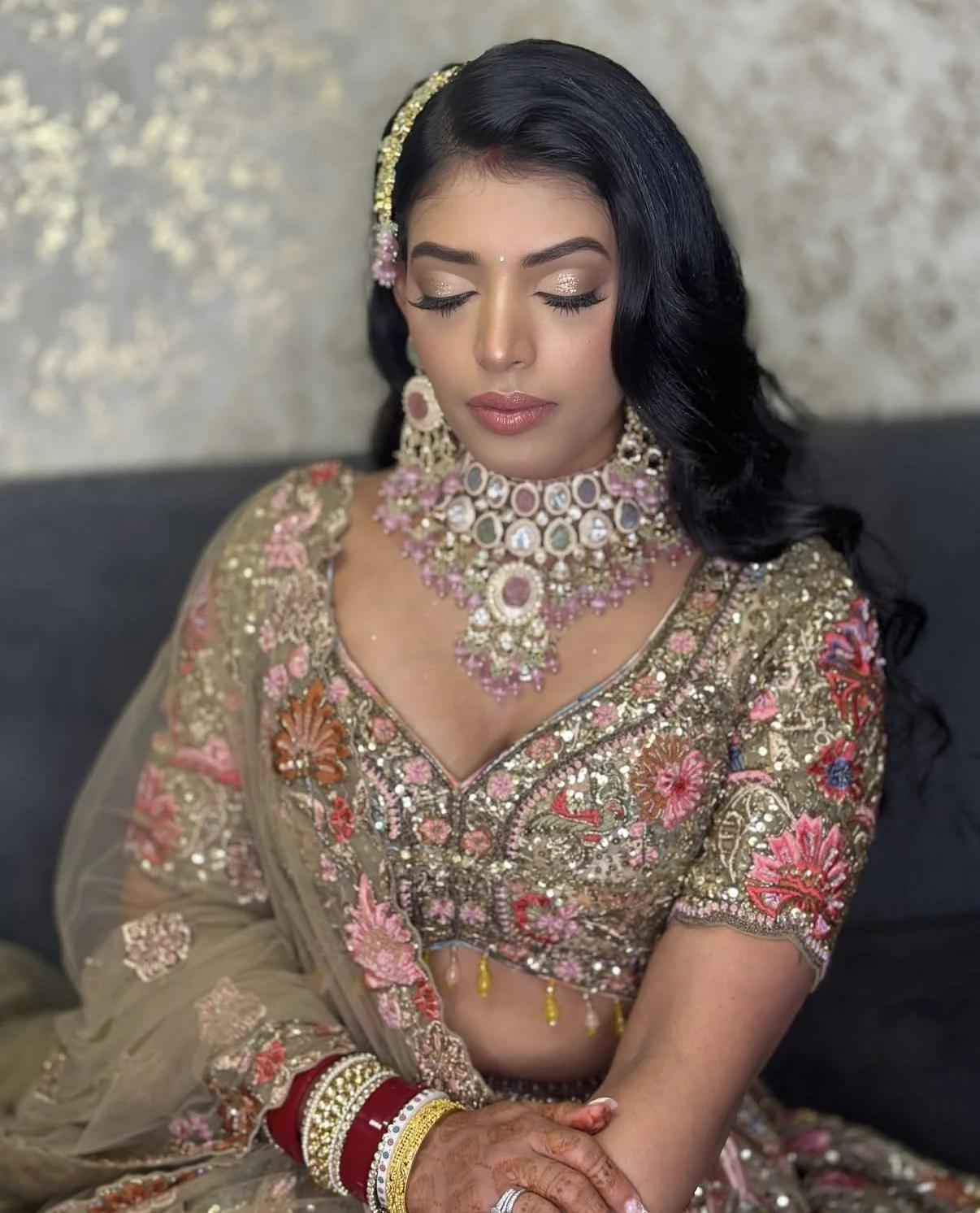 A woman dressed in traditional Indian bridal attire with intricate embroidery, heavy jewelry, and henna on her hands, sitting with her eyes closed.