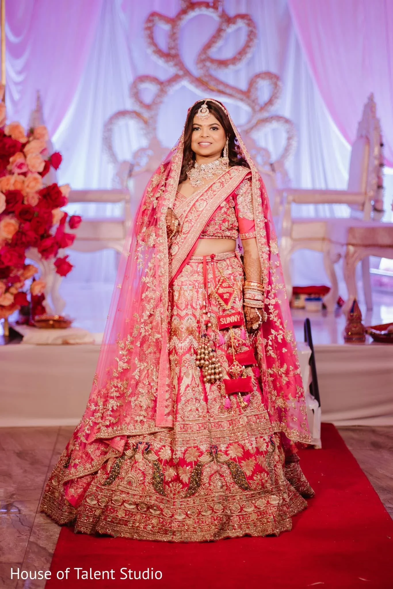 A woman dressed in a traditional ornate red and gold bridal lehenga, standing on a red carpet in an elegant decorated hall with pink and white drapes, floral arrangements, and ornate chairs, celebrating as a bride.