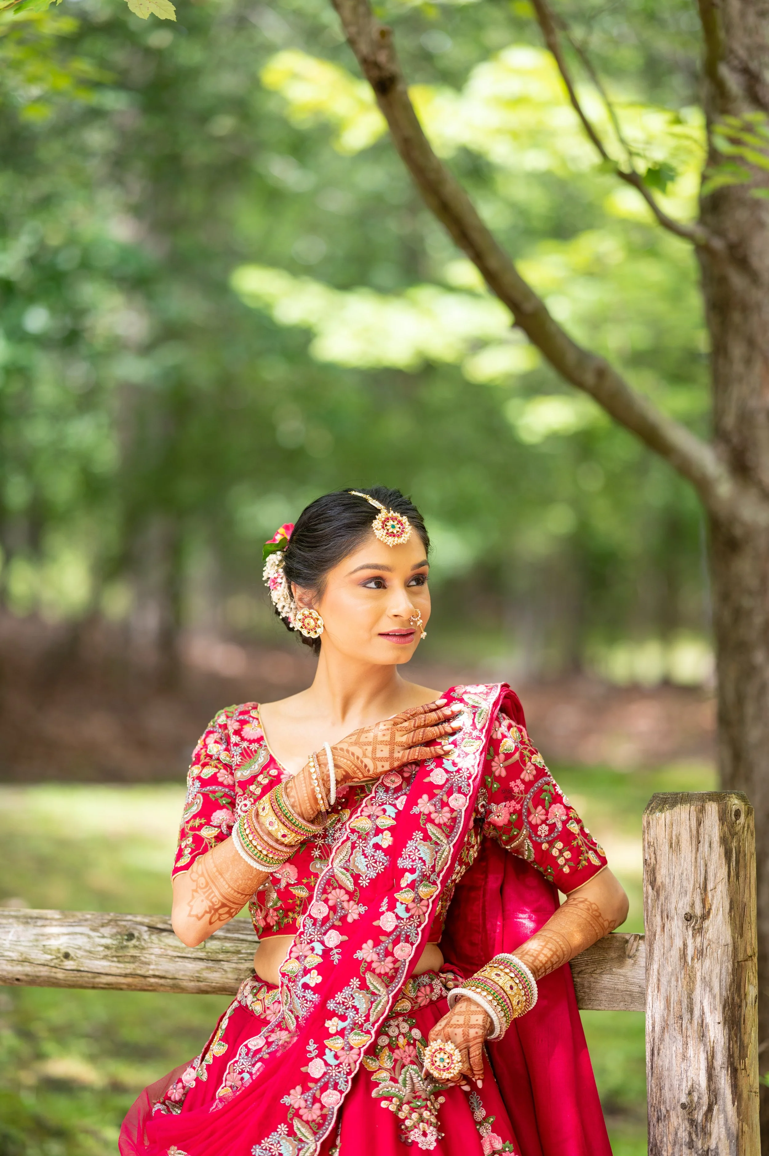 A woman dressed in traditional Indian attire with intricate jewelry and henna on her hands, standing outdoors among trees and greenery.