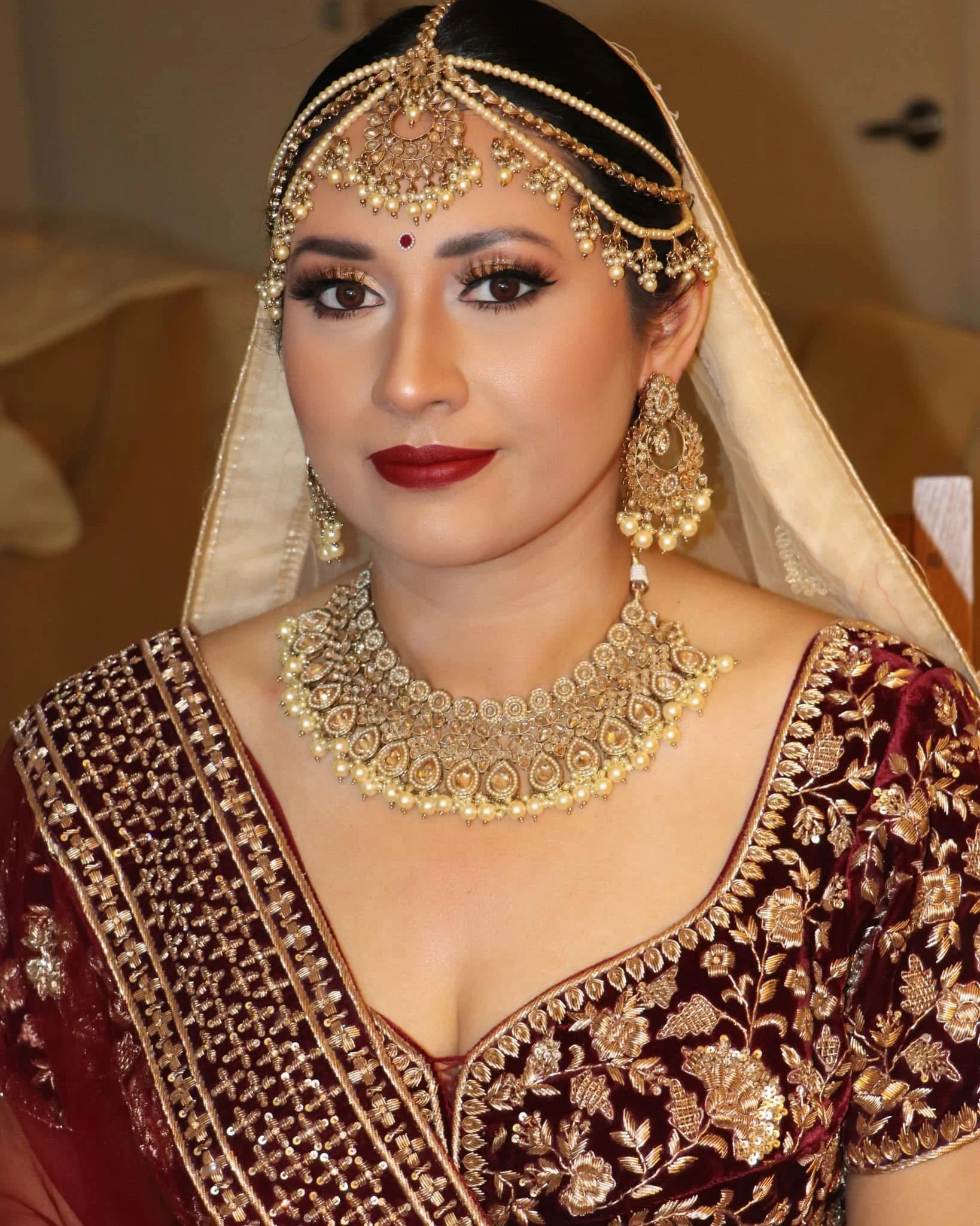 A woman dressed in traditional Indian bridal attire with gold jewelry including a necklace, earrings, and a headpiece, wearing a maroon and gold embroidered saree.