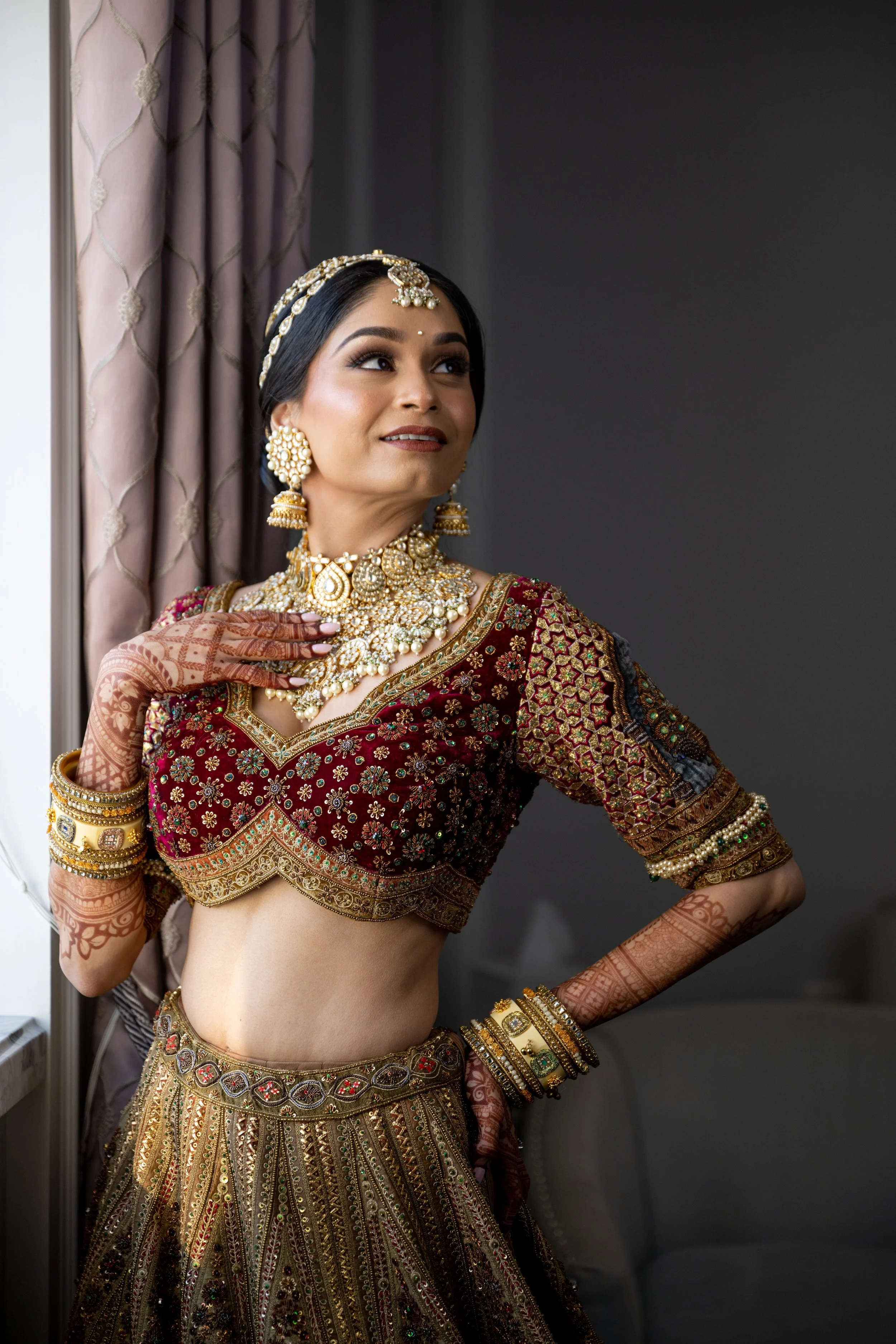 A woman dressed in traditional Indian bridal attire with ornate jewelry, henna on her hands and arms, standing near a window with purple curtains, looking up.