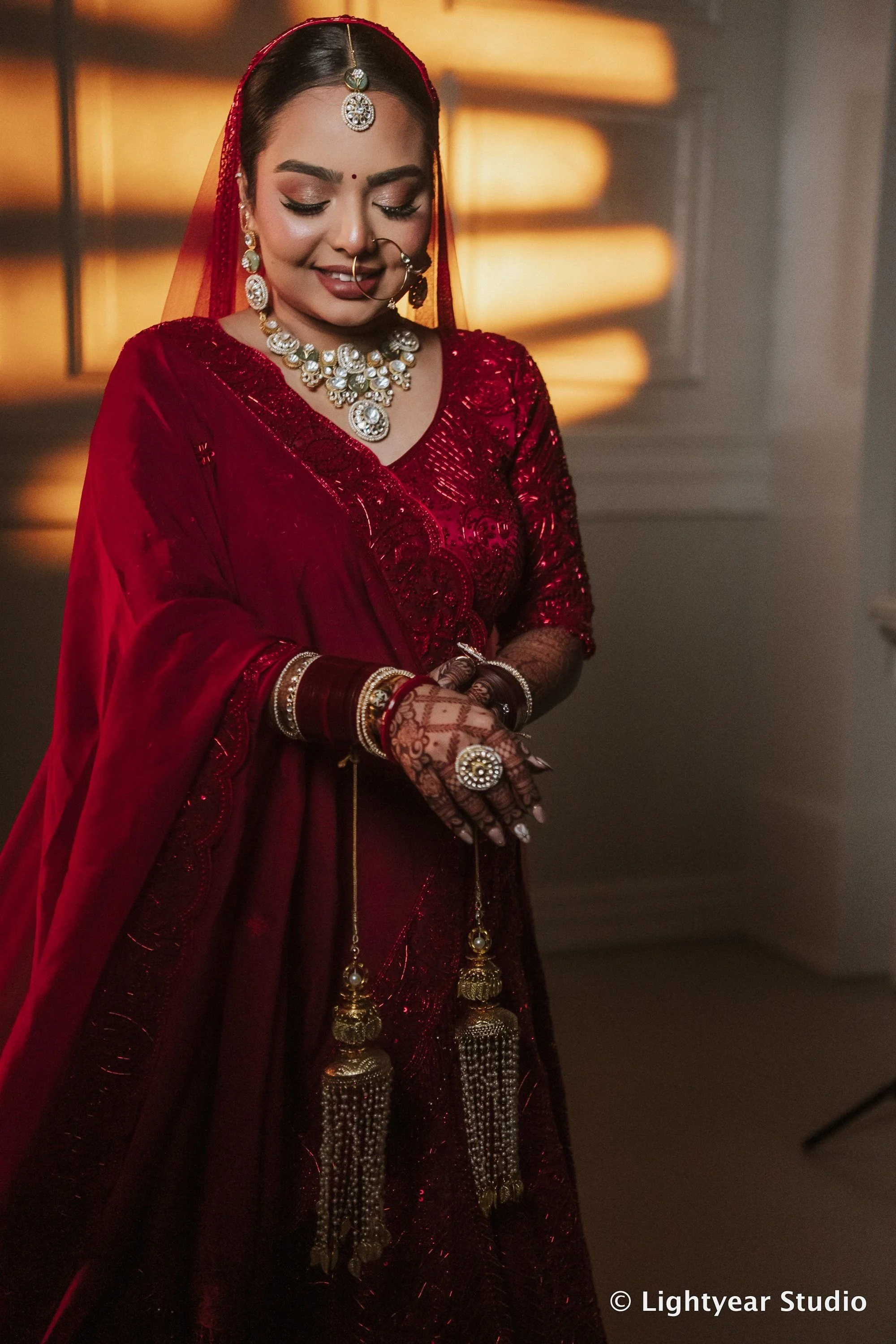 A woman dressed in traditional Indian attire, wearing a red embroidered saree with gold jewelry and henna on her hands, smiling with eyes closed in front of a warm-toned background.