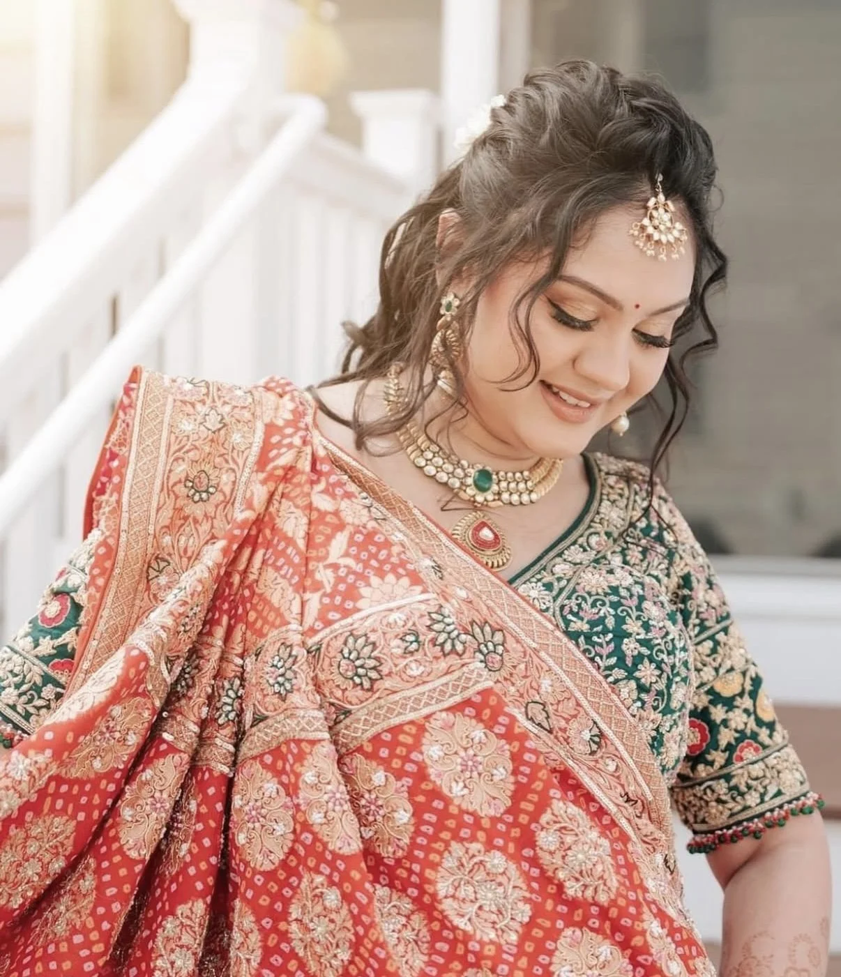 A woman dressed in traditional Indian attire with ornate jewelry, smiling and looking down.