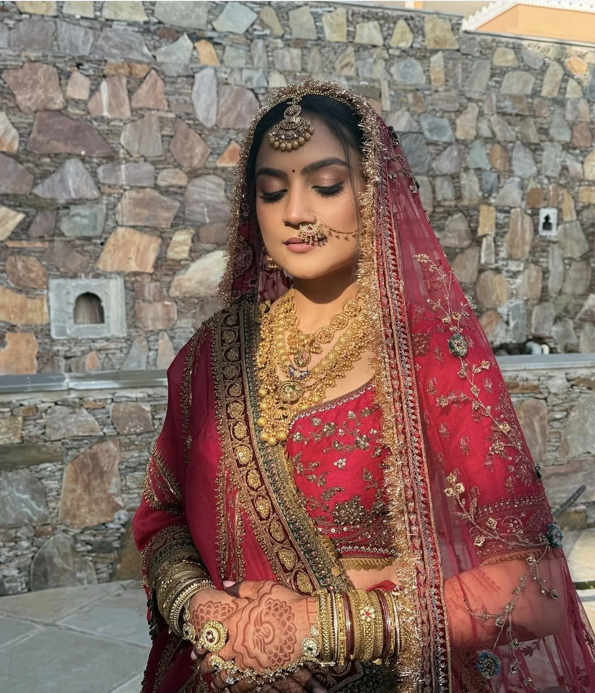 A woman dressed in traditional Indian bridal attire with red and gold embroidered saree, adorned with elaborate jewelry including necklaces, earrings, nose ring, and bangles, standing against a stone wall.