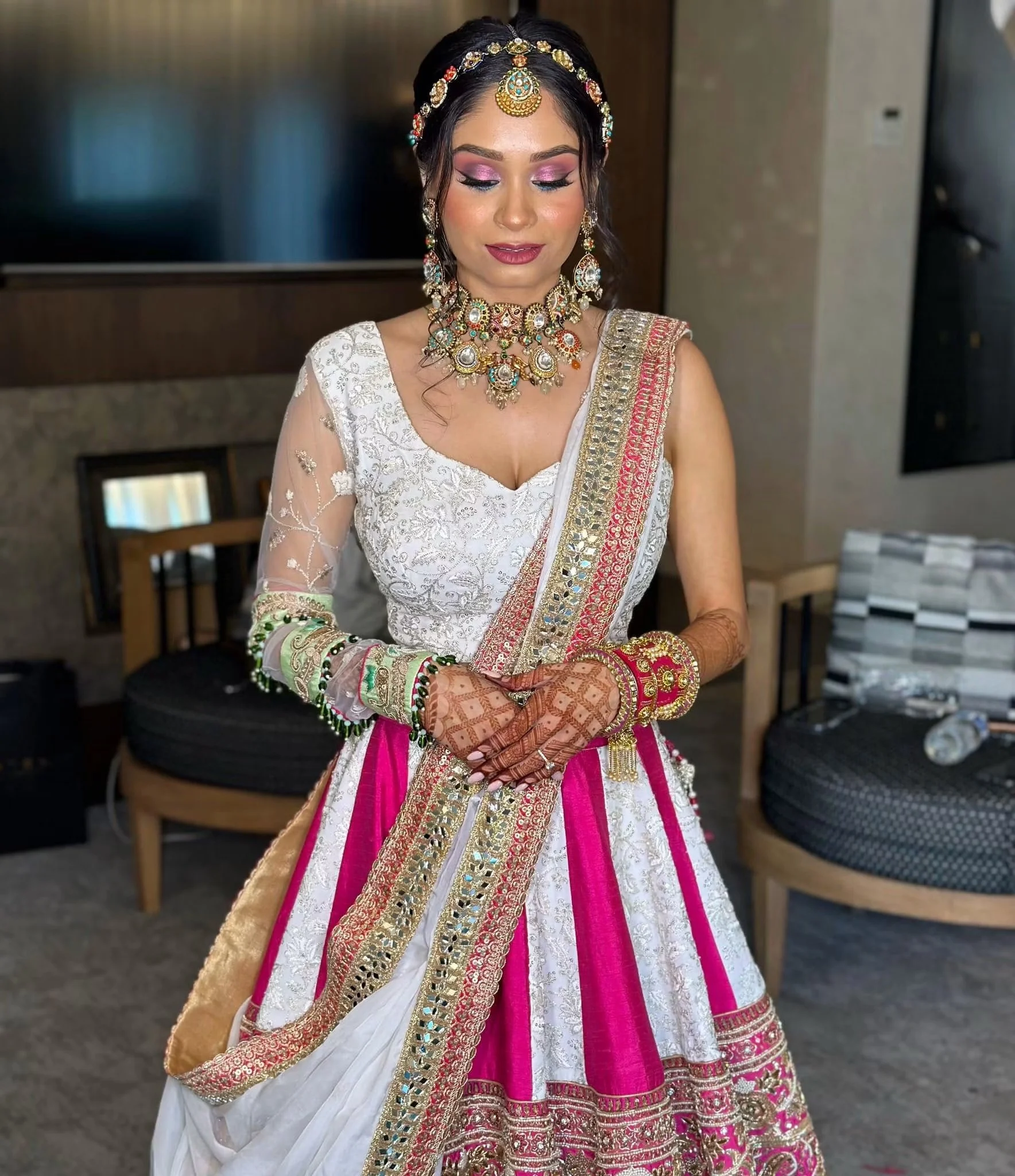 A woman dressed in a traditional Indian bridal outfit, wearing a white and pink lehenga with gold embroidery, and adorned with elaborate jewelry including a necklace, earrings, and a headpiece. She has henna on her hands and is standing indoors.