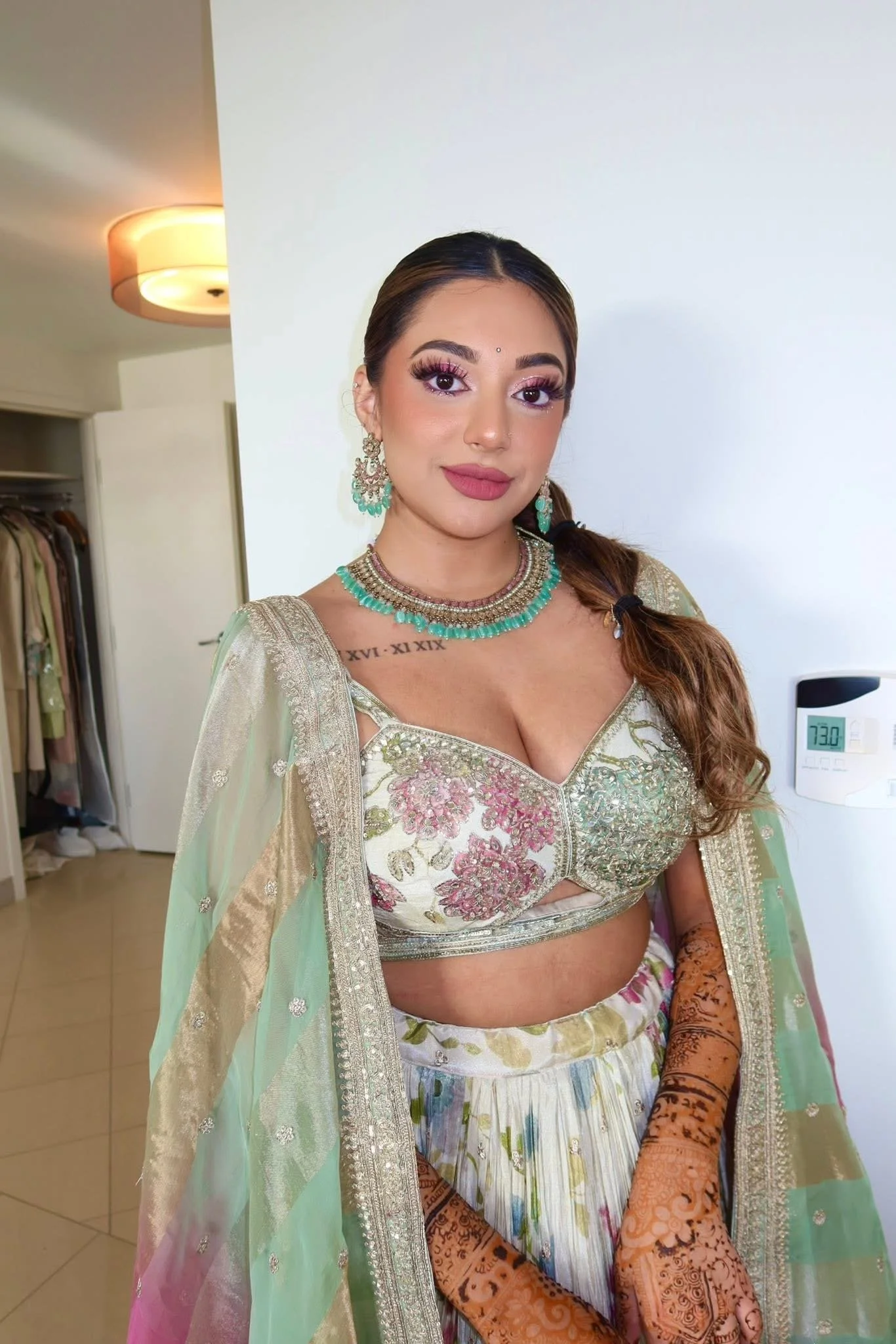 A woman dressed in traditional Indian attire with intricate Henna designs on her hands, wearing jewelry and makeup, standing indoors.