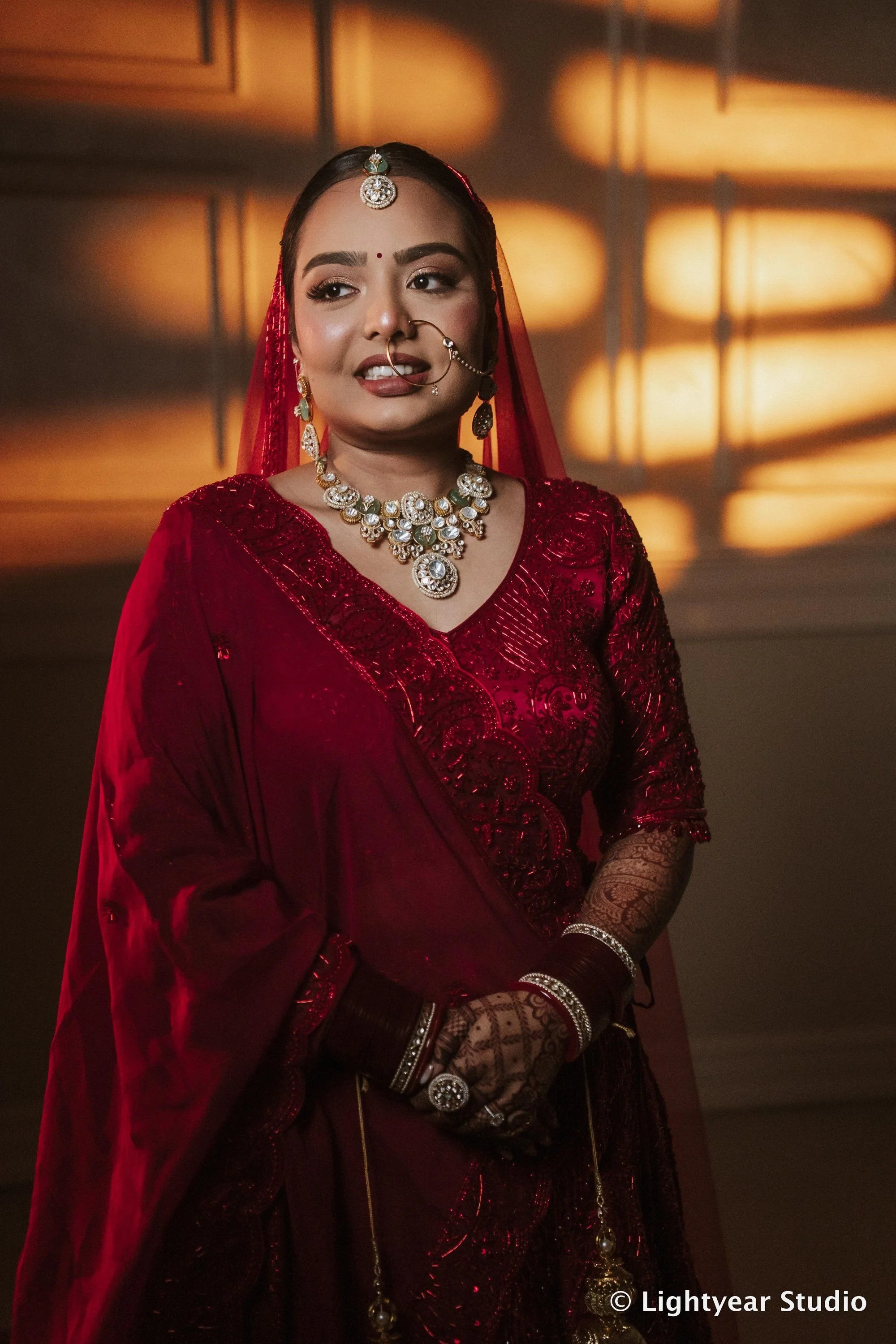 A woman of South Asian descent dressed in a rich red traditional outfit, adorned with jewelry including a necklace, earrings, and a headpiece, with intricate henna designs on her hands, standing against a warmly lit background.