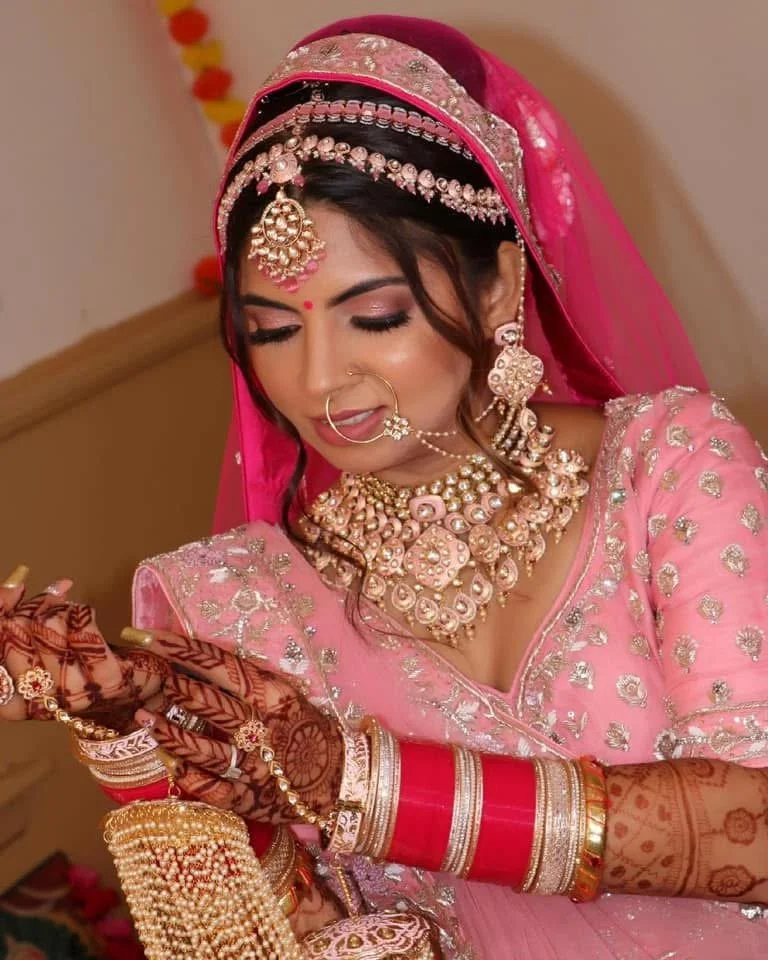 An Indian bride dressed in pink traditional attire, adorned with jewelry including a maang tikka, necklace, earrings, nose ring, and multiple bangles, looking at her phone.