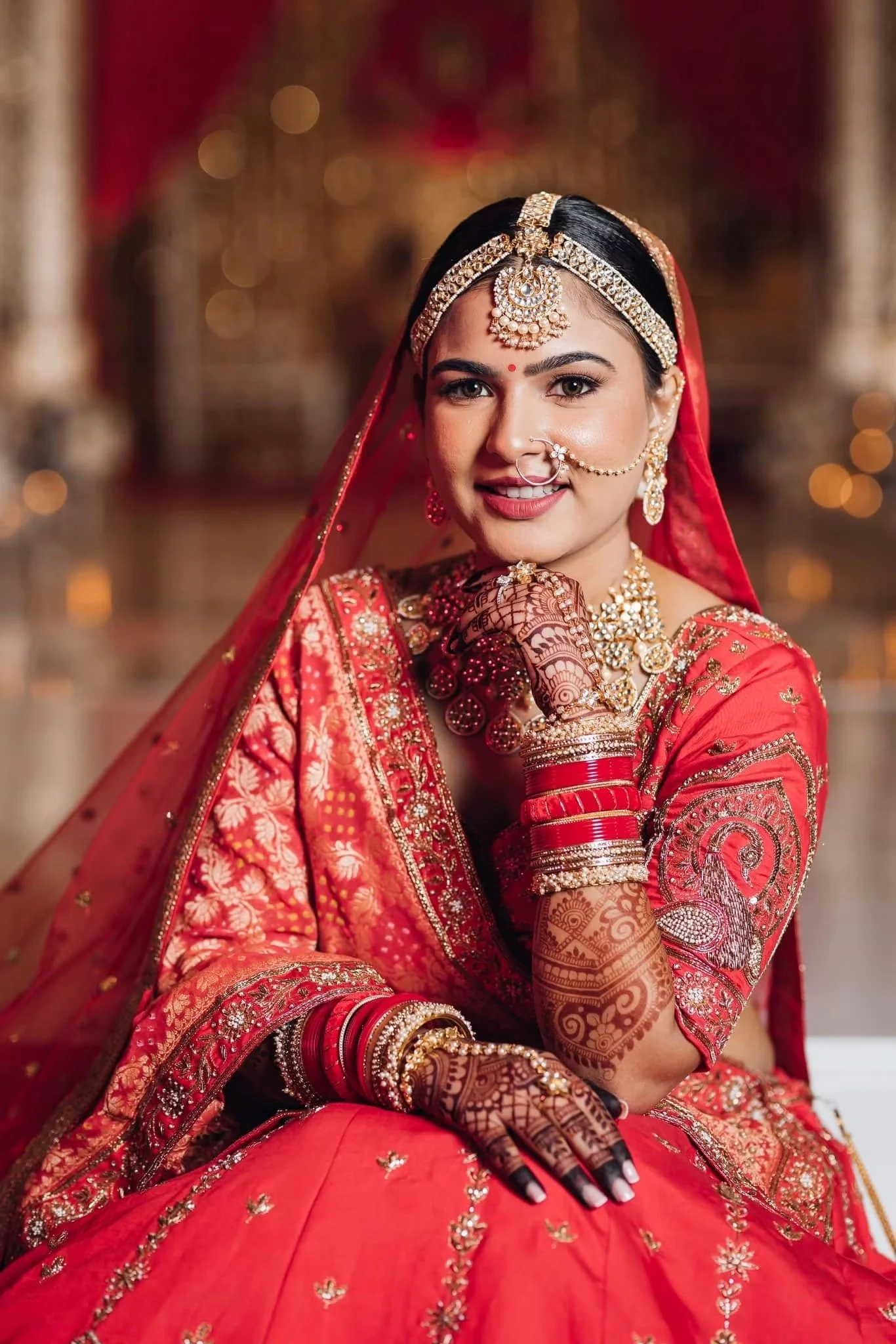 A woman dressed in traditional Indian bridal attire, wearing a red embroidered saree or lehenga, gold jewelry, a maang tikka on her forehead, a nose ring chain, and henna on her hands, smiling and looking at the camera.
