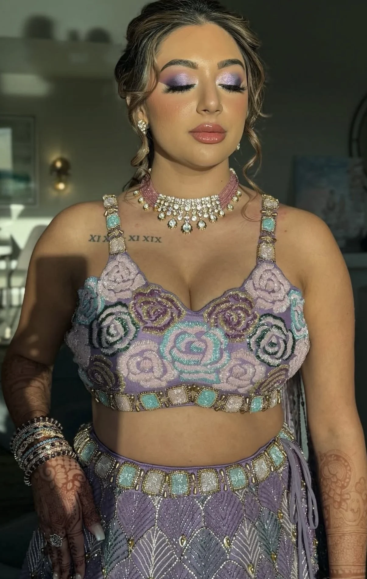 A woman with closed eyes wearing purple eyeshadow and glossy lips. She is dressed in a lavender embroidered outfit with pastel and gold accents, adorned with jewelry including a necklace, earrings, and bracelets, and has henna designs on her hands an