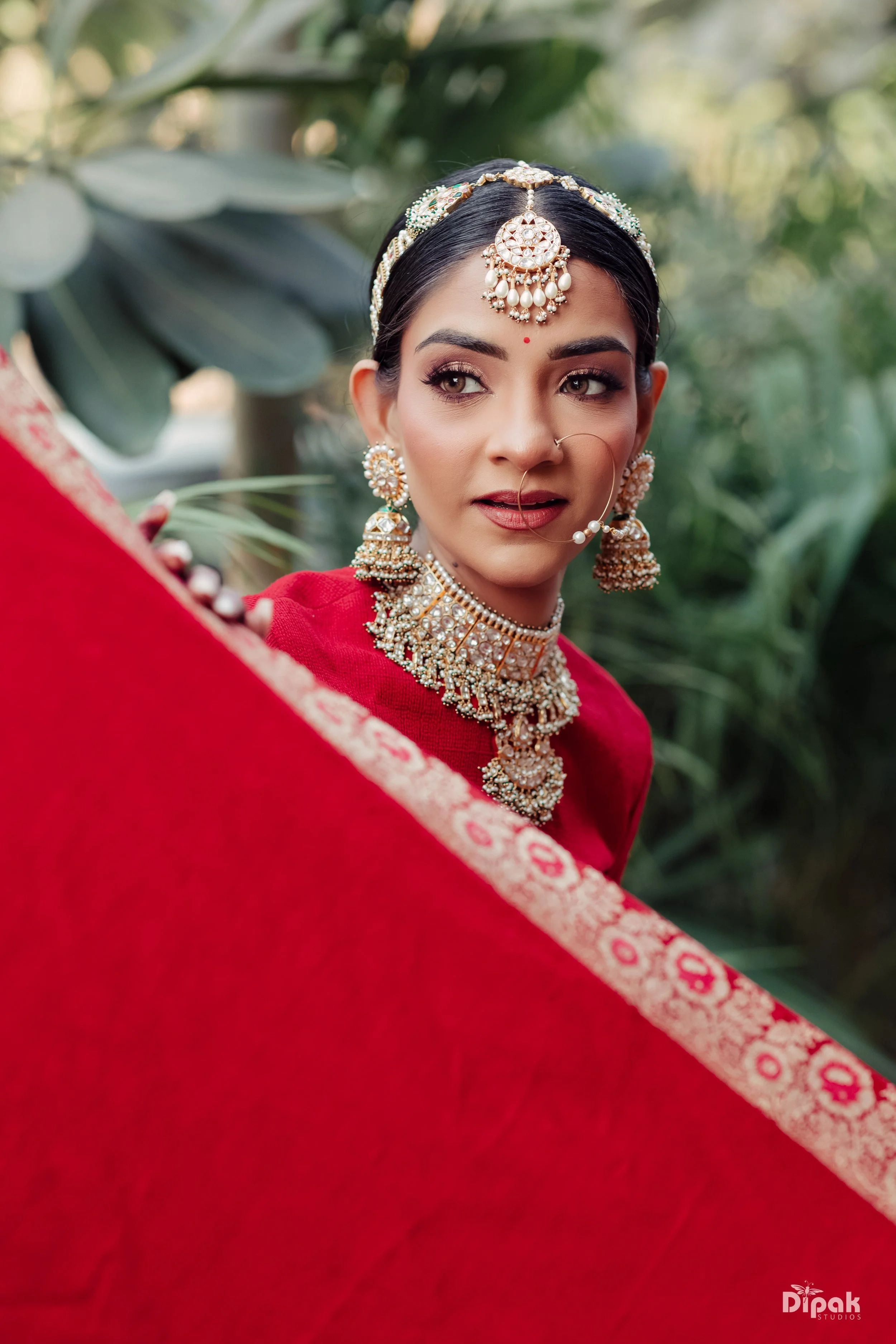 A woman dressed in traditional Indian attire adorned with elaborate gold and pearl jewelry, including a necklace, earrings, and a headpiece, seen outdoors with greenery in the background.