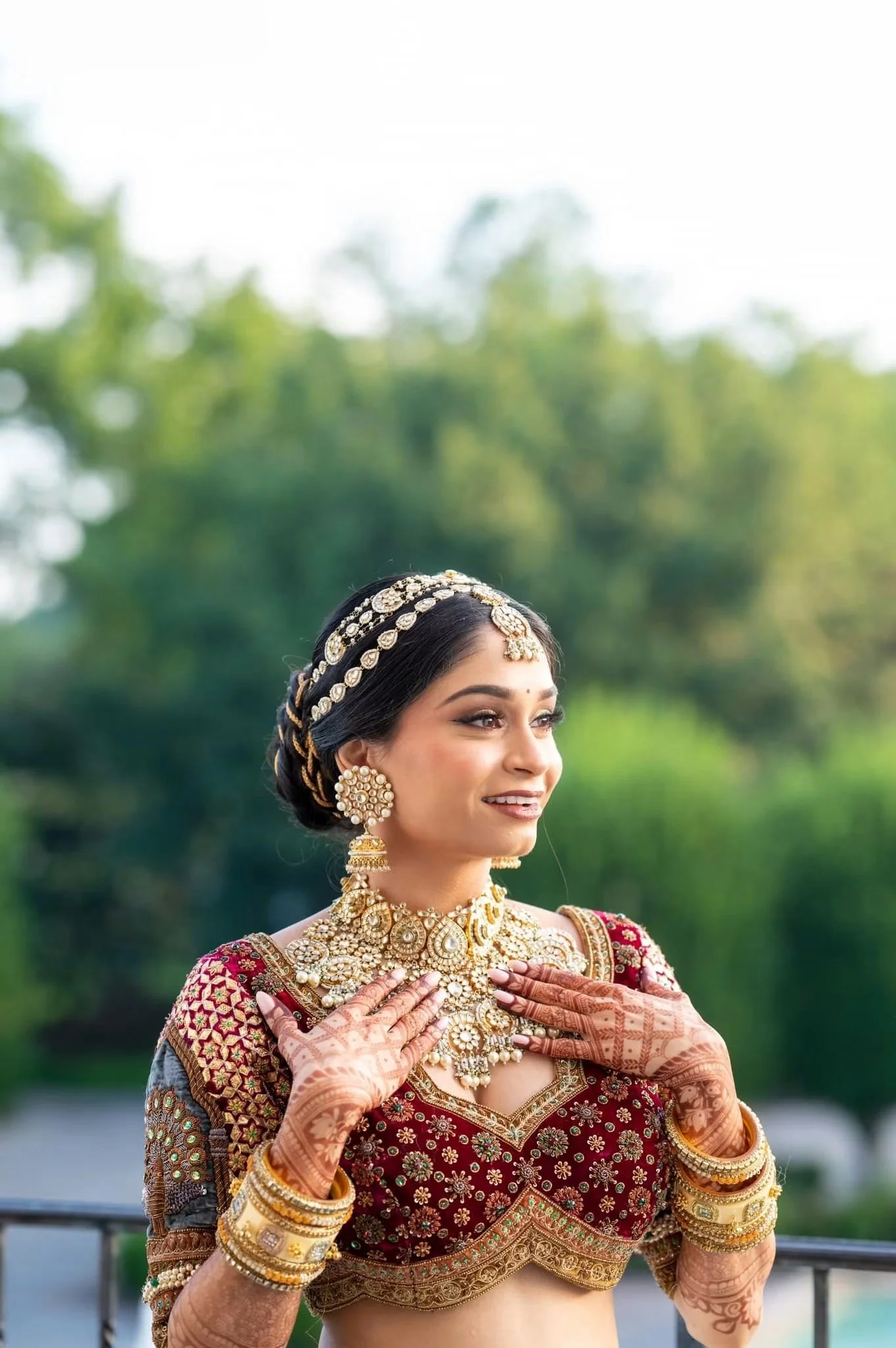 A woman wearing traditional Indian jewelry and attire, holding her hands near her chest with henna designs, outdoors with greenery in the background.