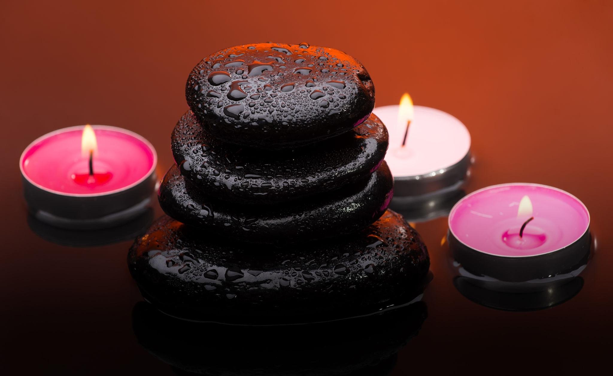 Stack of wet black spa stones surrounded by three lit pink candles on a reflective surface with a warm orange background.