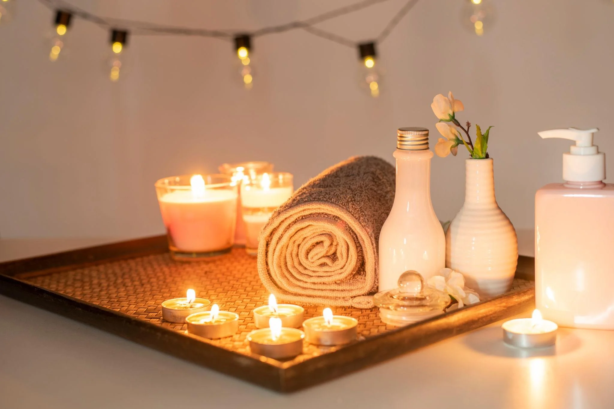 Spa setting with lit candles, rolled towel, and bottles with flowers, creating a relaxing atmosphere.