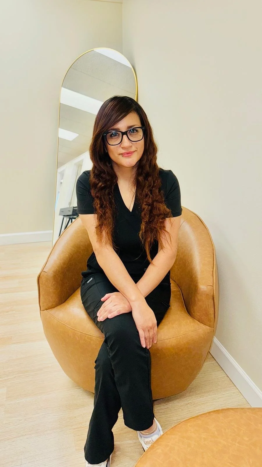 A woman with long, wavy brown hair, wearing glasses and a black outfit, sitting on a tan, rounded chair in a room with light-colored walls.