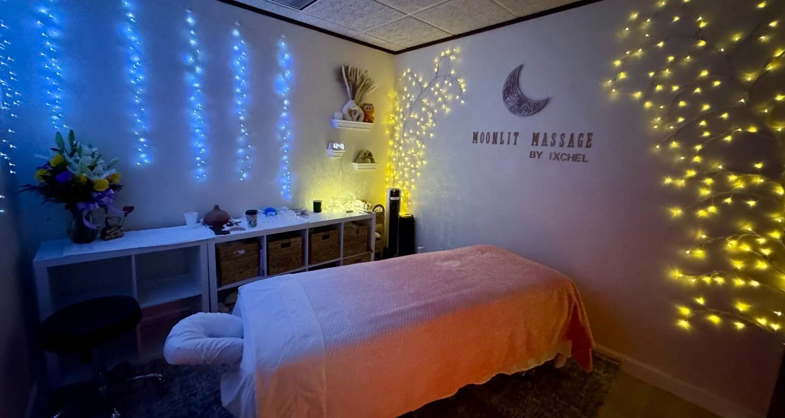 A massage room with blue and yellow string lights, a massage table with an orange cover, a white shelf with decorative items, and wall art that reads 'Moonlit Massage by Xchel'.