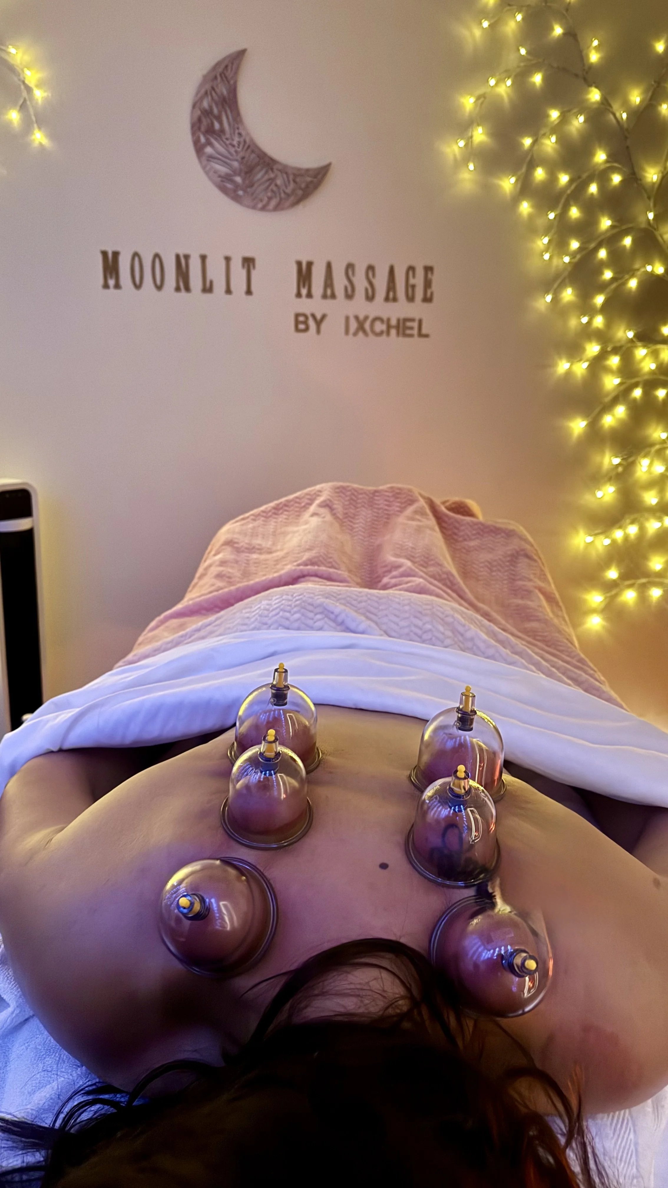 Person receiving a cupping therapy session, lying face down on a treatment table with glass cups placed on their back, in a room with decorative lights and a sign that reads 'Moonlit Massage by Ixchel'.