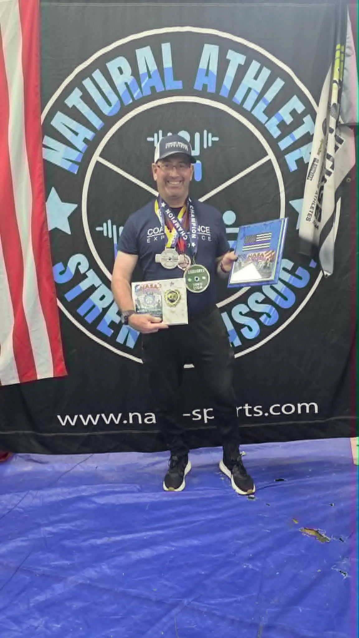 Man standing on a blue gym floor, smiling, holding medals and a certificate, in front of a 'Natural Athletes' banner, with a flag and hockey stick nearby.
