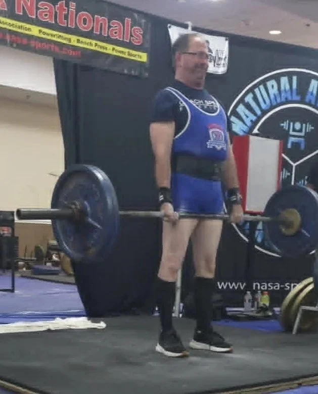 A man lifting a barbell during a powerlifting competition, wearing a singlet, knee-high socks, and wrist supports, standing on a lifting platform. He appears to be ready to perform a deadlift.