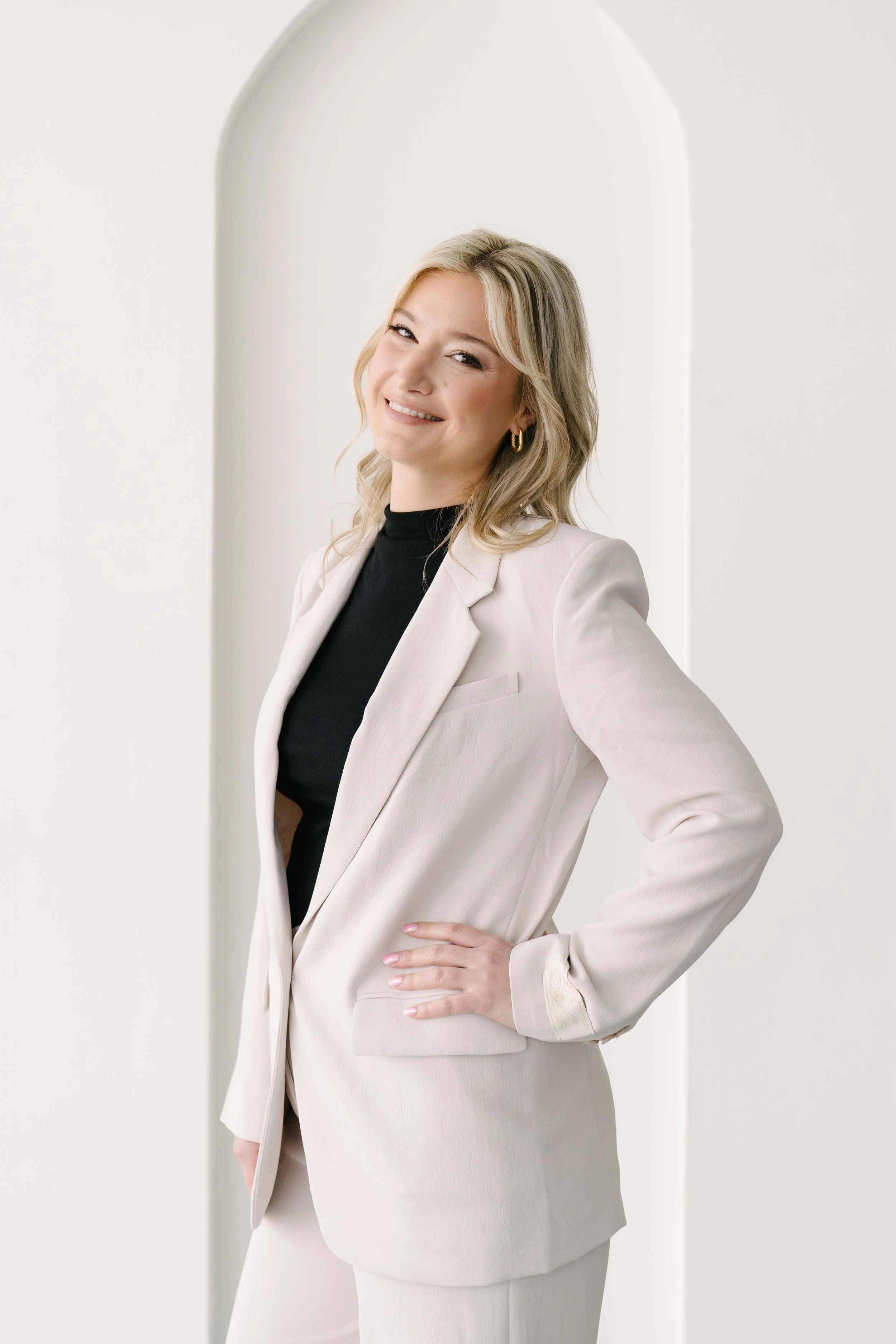 A woman with blonde hair, wearing a white blazer and black top, smiling and posing with one hand on her hip against a white background with an arched architectural feature.