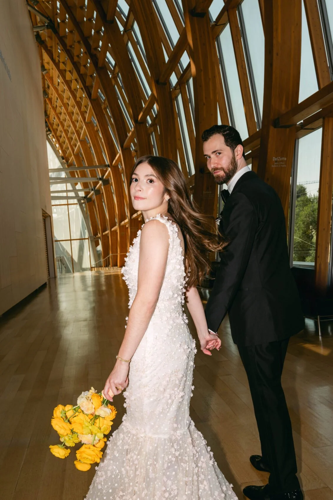 A bride and groom holding hands inside a modern wooden building with large windows. The bride wears a floral lace wedding gown and holds a bouquet of yellow flowers. The groom wears a black tuxedo with a bow tie.