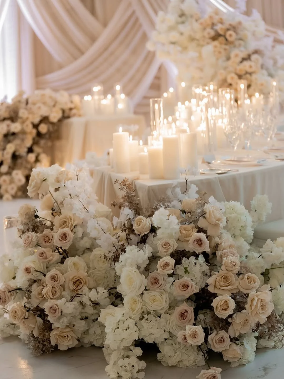 Elegant wedding table decorated with white and blush roses, white hydrangeas, candles, and glassware with a backdrop of draped fabric and flowers.