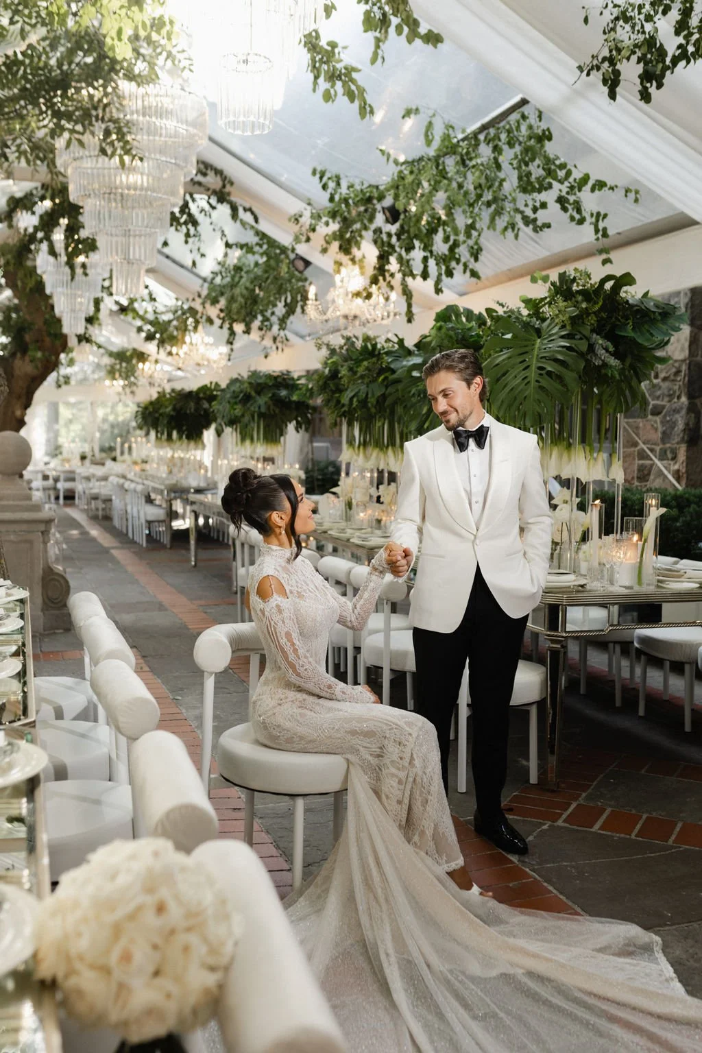 A bride and groom at their wedding reception holding hands and smiling at each other in an elegant, decorated venue with greenery and chandeliers.