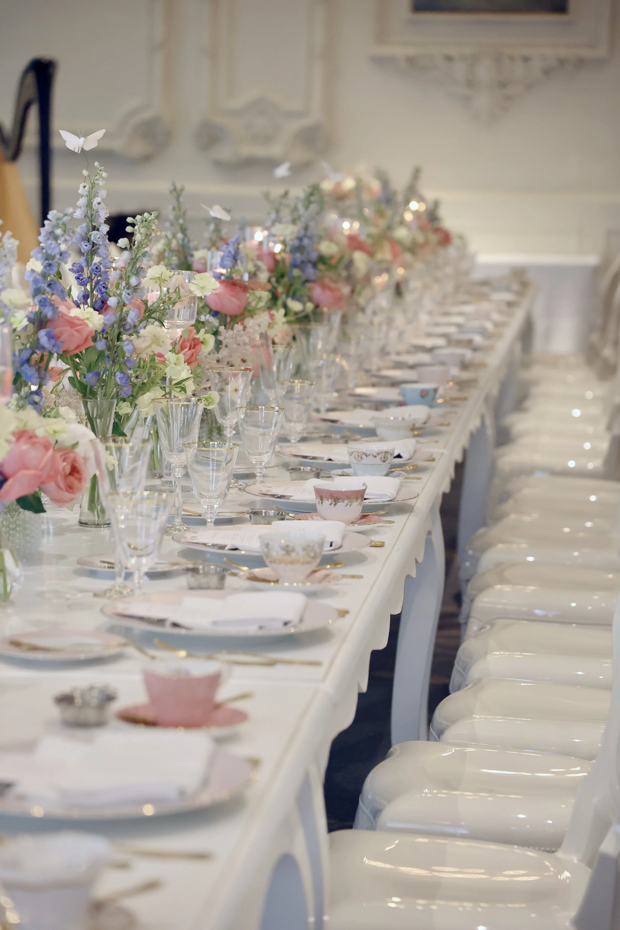A long banquet table decorated with pastel-colored flowers, glassware, and fine china, set for a formal event in an elegant room.