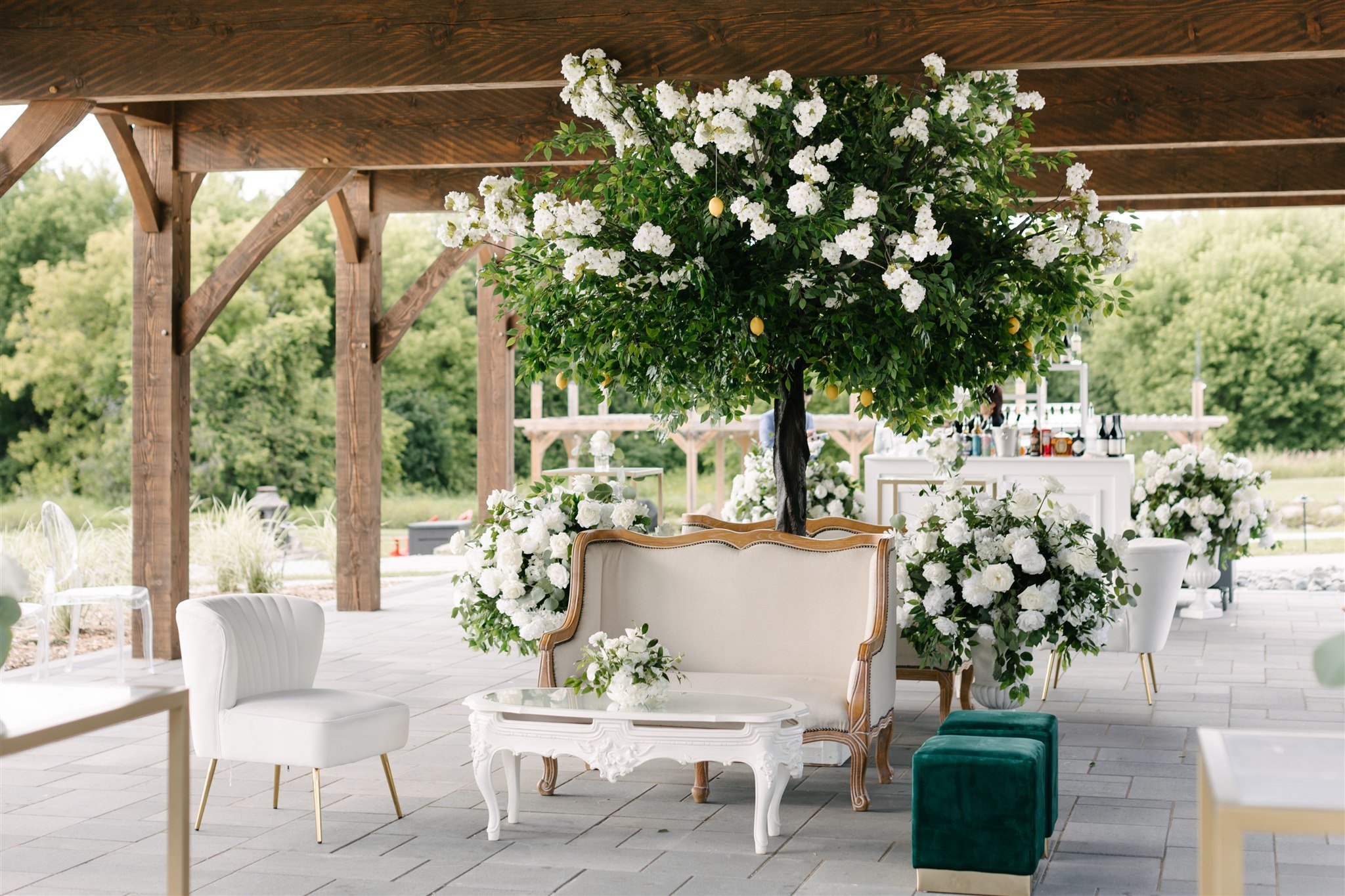 Outdoor event setup with a large tree decorated with white flowers and mini lemons, surrounded by white lounge furniture and decor under a wooden pavilion with greenery in the background.
