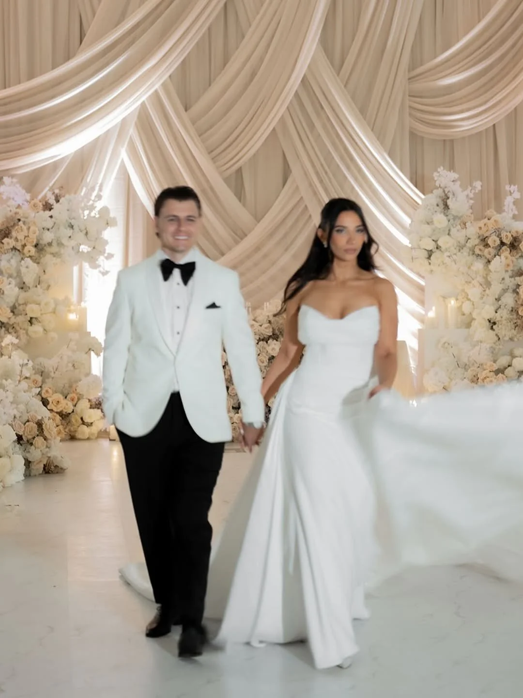 A newlywed couple walking down the aisle, holding hands, in a decorated wedding venue with floral arrangements and draped fabric backdrop.