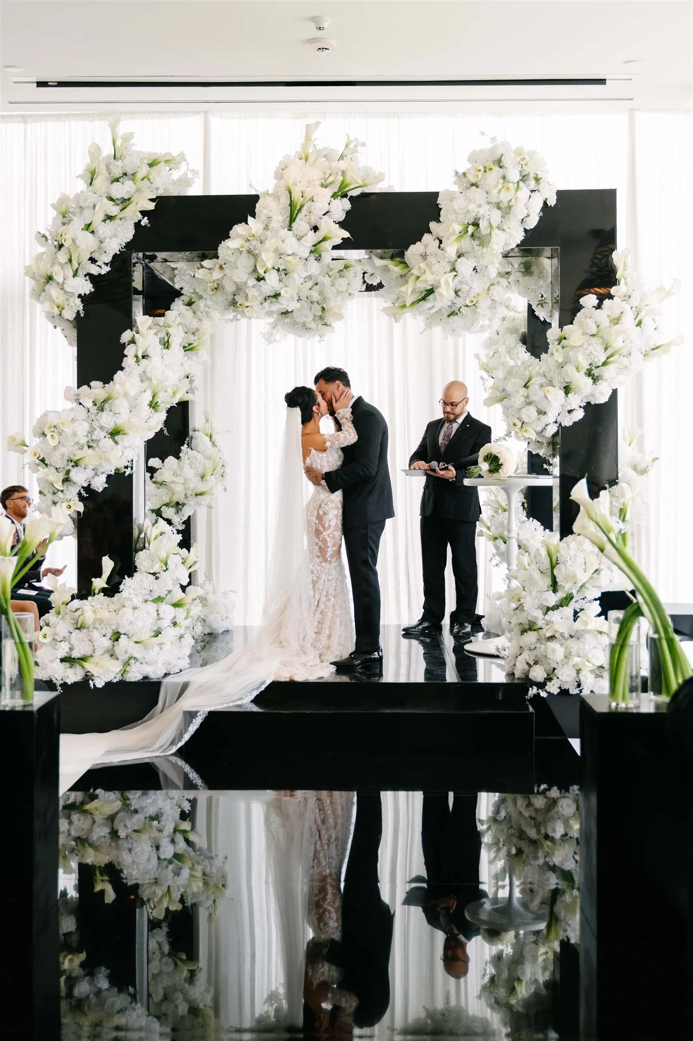 A wedding ceremony with a bride and groom kissing under a floral arch with white flowers, with a minister officiating and guests seated nearby.