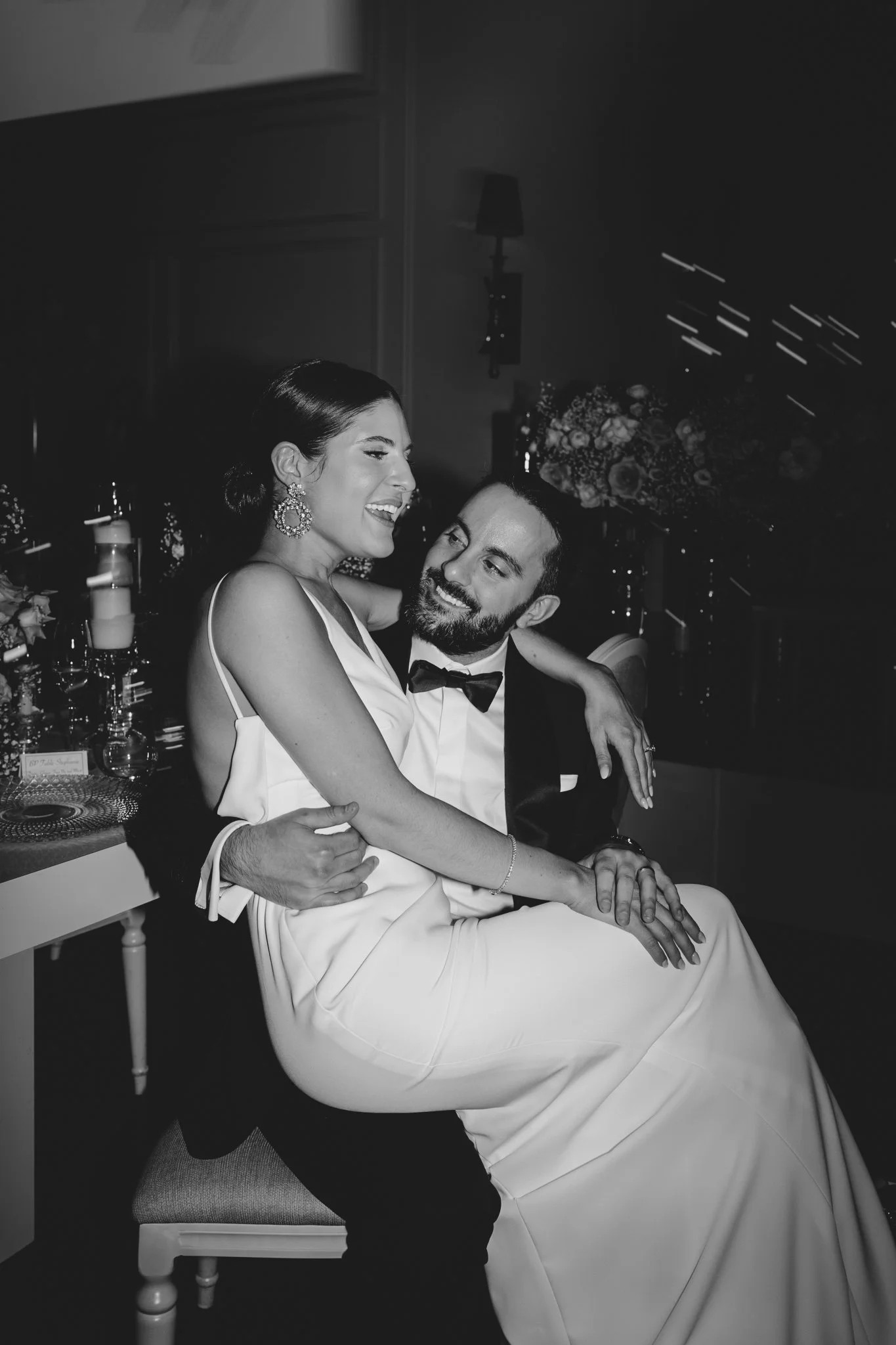 A couple dressed in formal attire, a woman in a white gown with large earrings and a man in a tuxedo, are sitting together at the wedding reception, smiling and sharing a joyful moment.