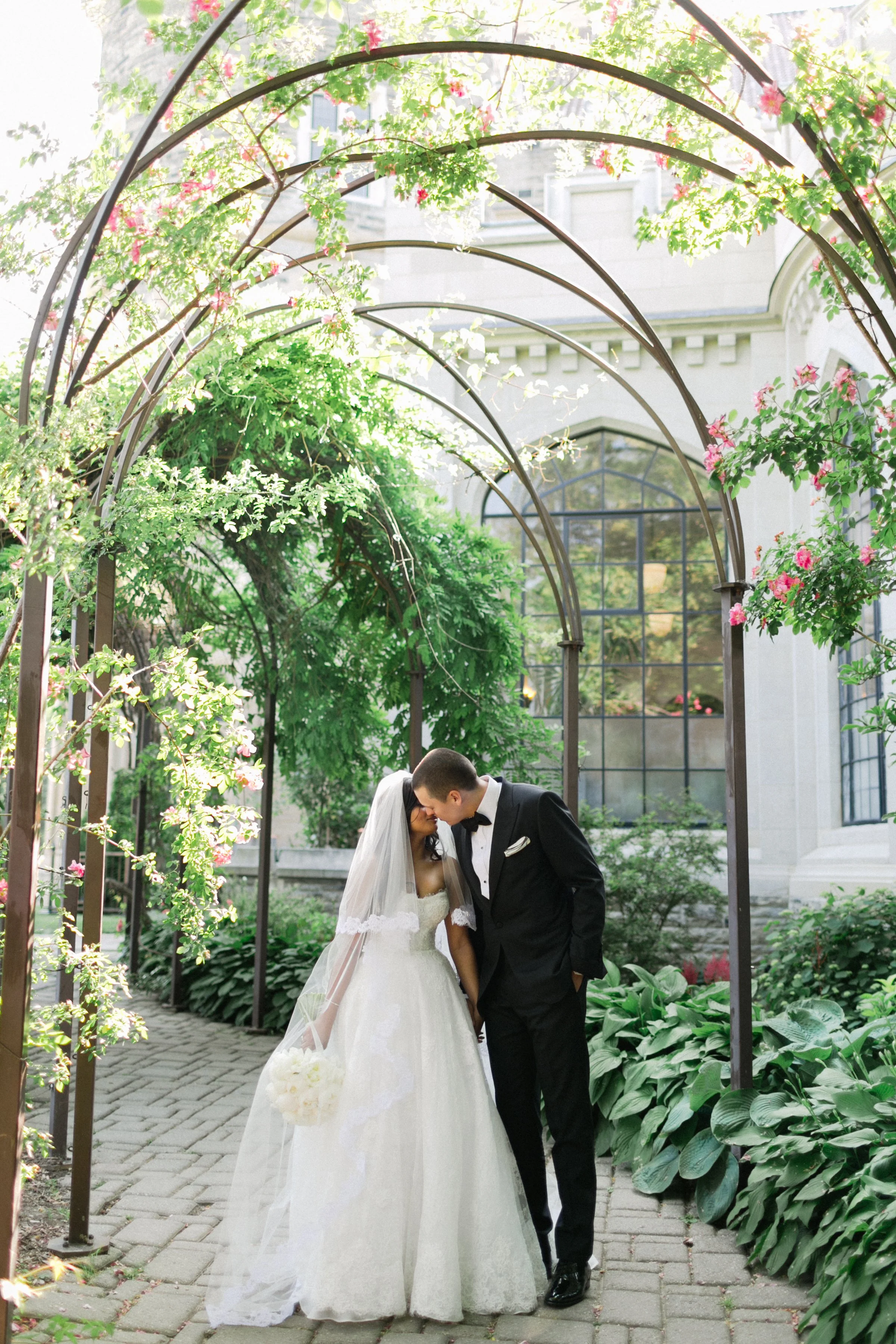 A bride and groom share a kiss under a flower-covered archway in a garden.