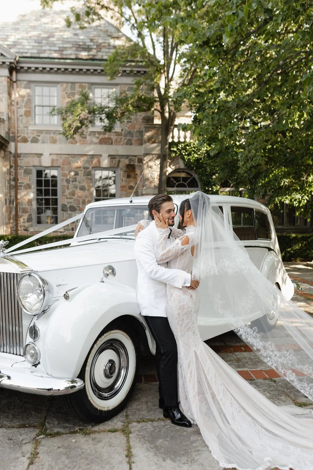 A bride and groom standing beside a vintage white car, sharing a romantic moment outdoors with a brick building and green trees in the background.