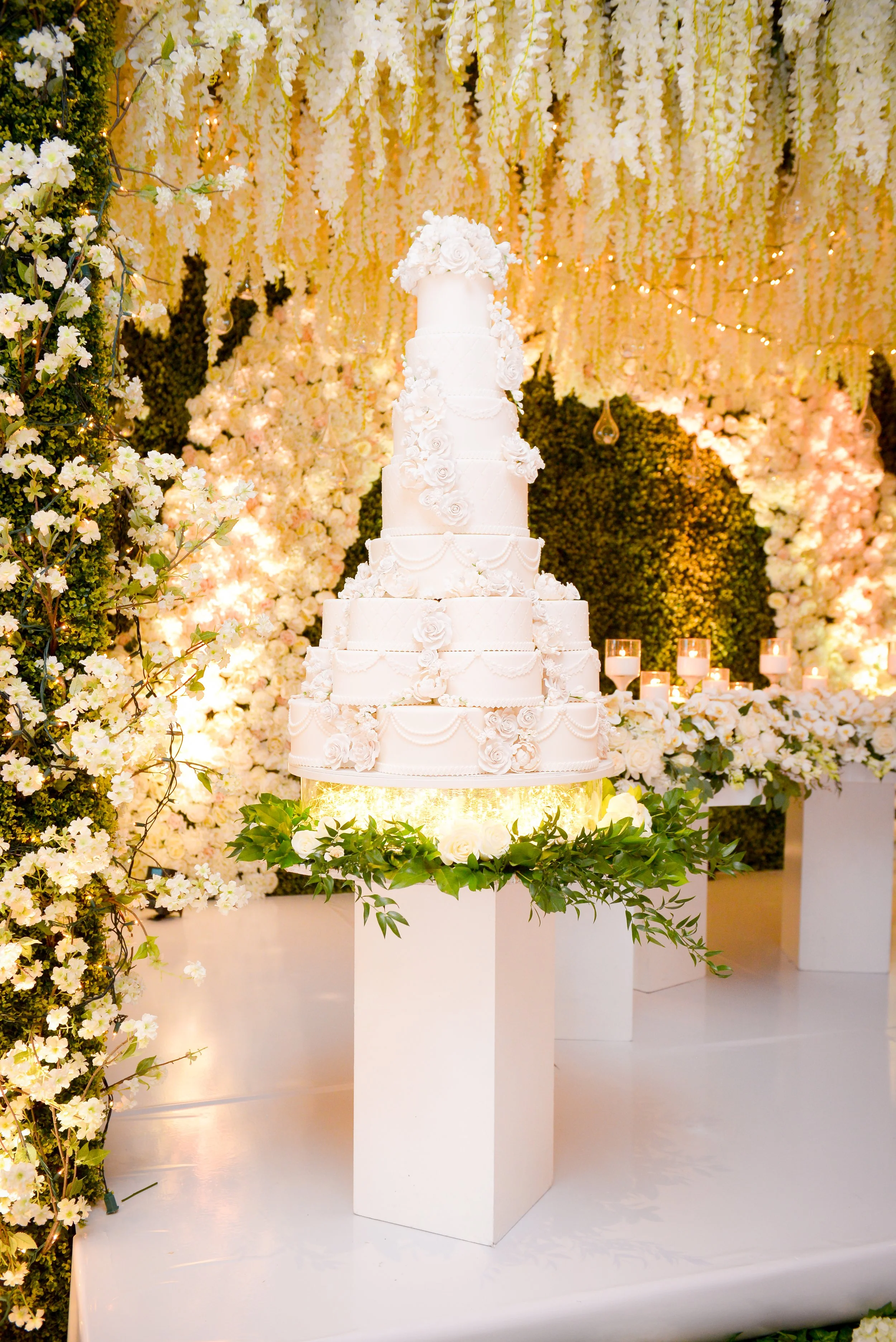 A multi-tiered white wedding cake decorated with white roses and floral accents, displayed on a round table with greenery. The cake is set against a backdrop of cascading white flowers, hanging lights, and candles, creating an elegant wedding decorat