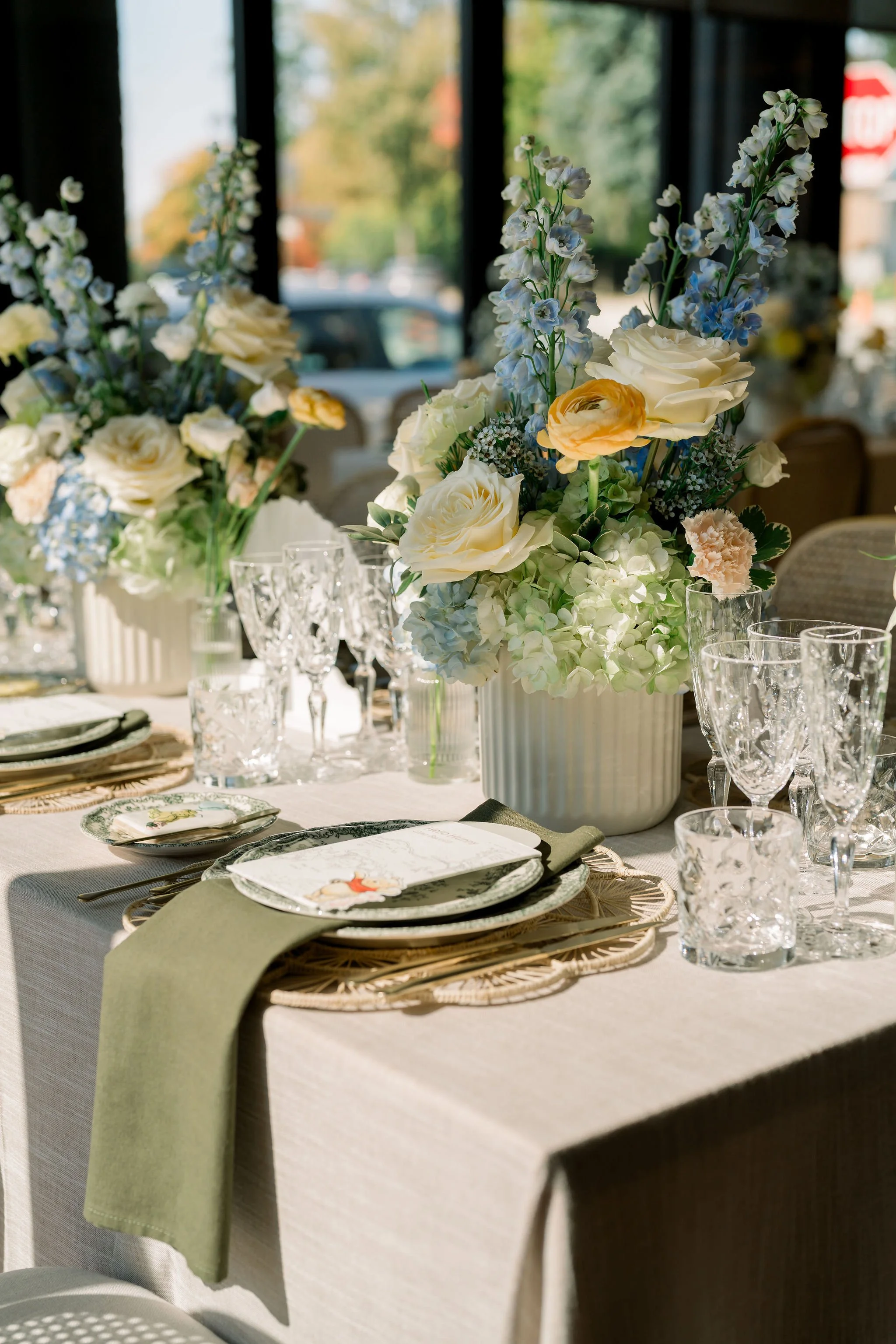 Elegant dining table decorated with a large floral centerpiece featuring white roses, blue delphiniums, and green foliage in a white ribbed vase; table setting includes patterned plates, a menu card, crystal glassware, and silverware, with a light gr
