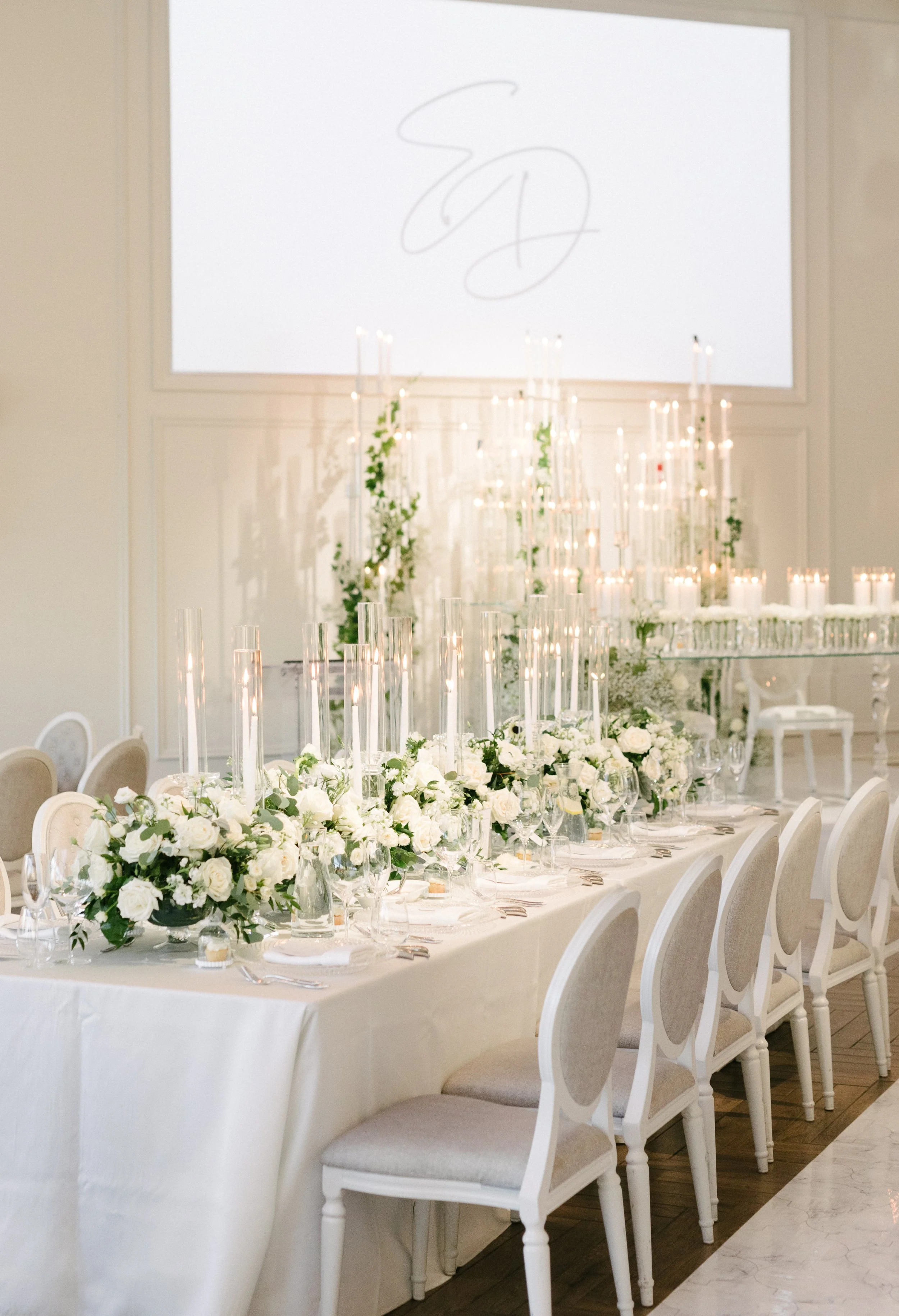 Elegant banquet table set with white flowers, candles, and glassware. Surrounded by white chairs with beige cushioned seats, in a decorated event hall with a large white screen displaying initials.
